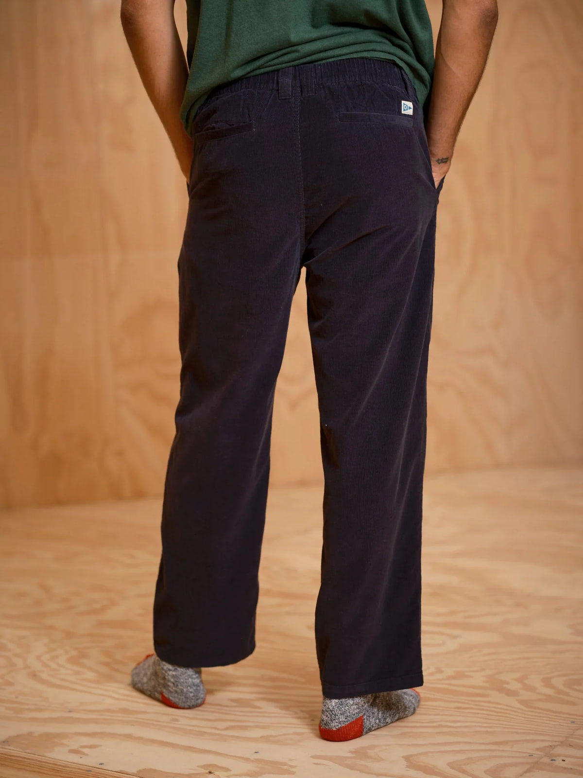 Back view from the waist down of a man wearing the Mollusk Boat Pants in Black Corduroy, showing the elastic back waistband with a small woven Mollusk label, two back welt pockets, relaxed straight-leg cut, and deep black micro-corduroy fabric, styled with a green tee against a plywood backdrop
