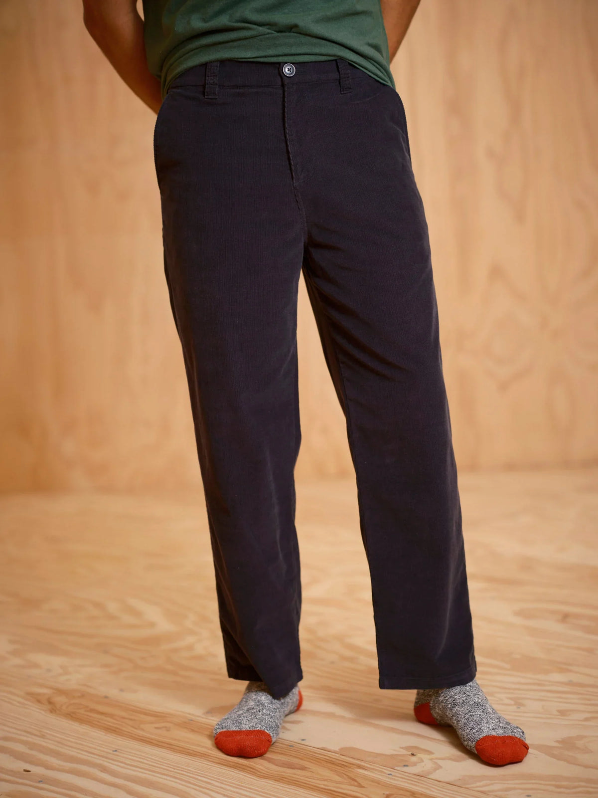 Front view from the waist down of a man wearing the Mollusk Surf Shop Boat Pants in Black Corduroy, showing classic chino-style structured front, zip fly with dark button closure, two front slash pockets, and relaxed straight-leg silhouette in deep black 100% cotton micro-corduroy, styled with a green tee and marled socks on a plywood floor

