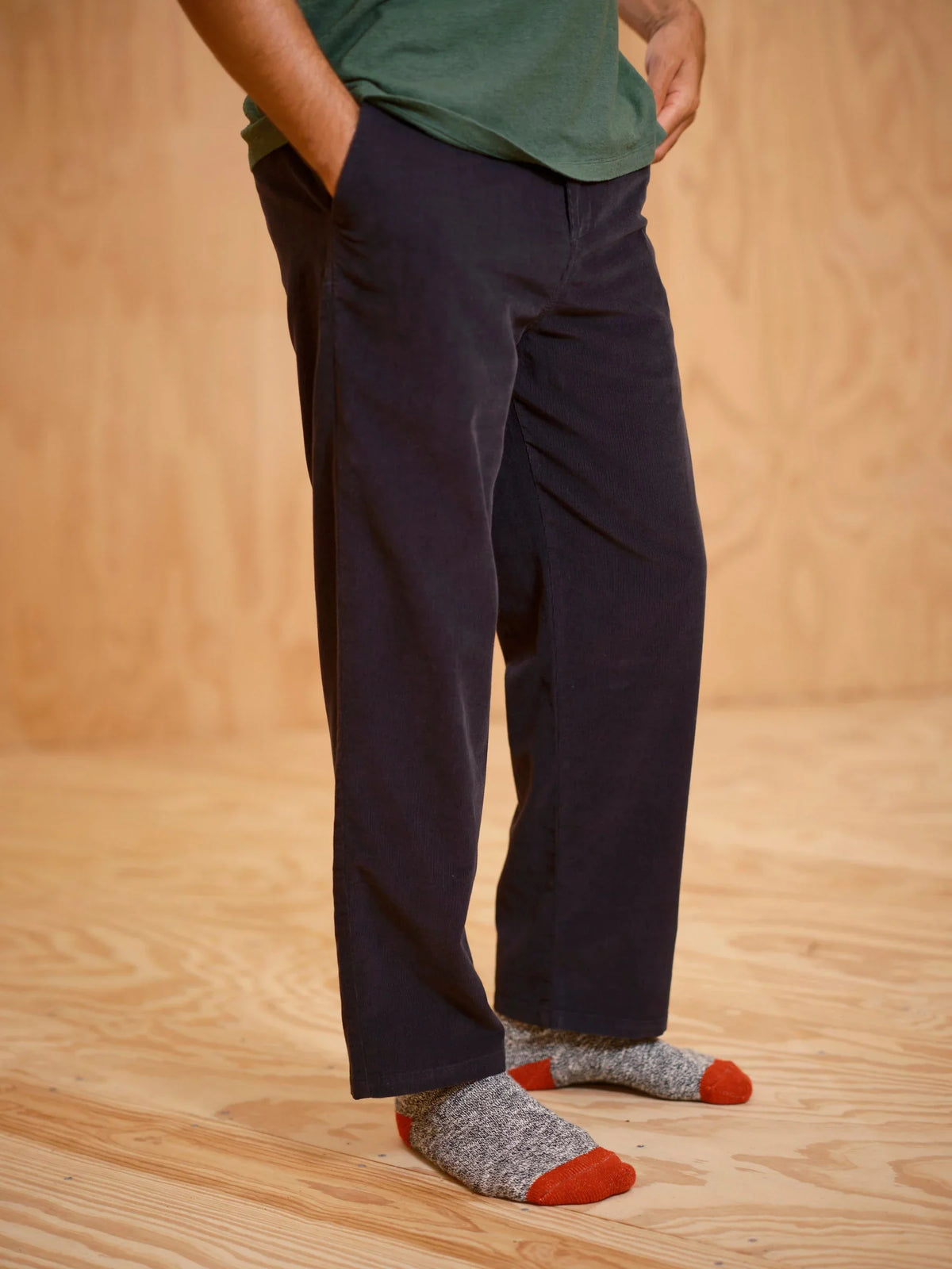 Side view from the waist down of a man wearing the Mollusk Boat Pants in Black Corduroy with hand in front pocket, showing the relaxed straight-leg drape, side-seam pocket opening, and soft micro-corduroy texture in deep black 100% cotton, styled with a green tee against a warm plywood backdrop
