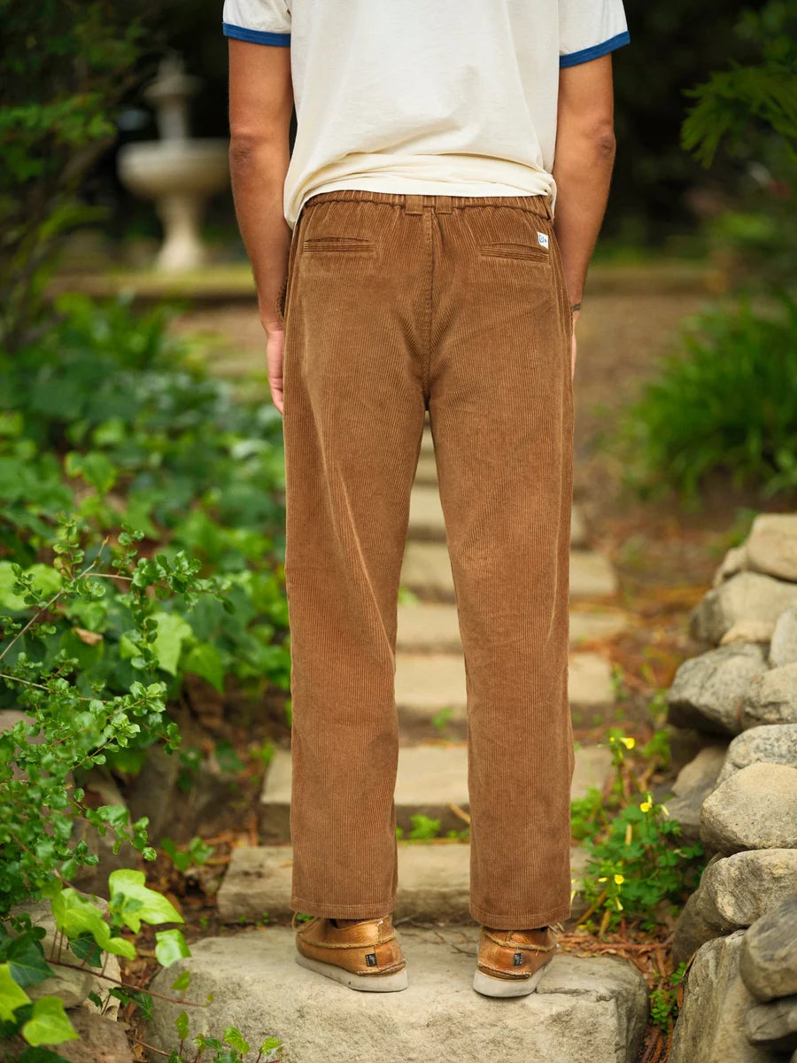 Back view from the waist down of a man wearing the Mollusk Boat Pants in Brown Wide Wale Corduroy, showing the elastic back waistband with a small woven label, two back pockets, relaxed straight-leg silhouette, and brown medium-wale corduroy, styled with a white tee and tan sneakers on stone garden steps

