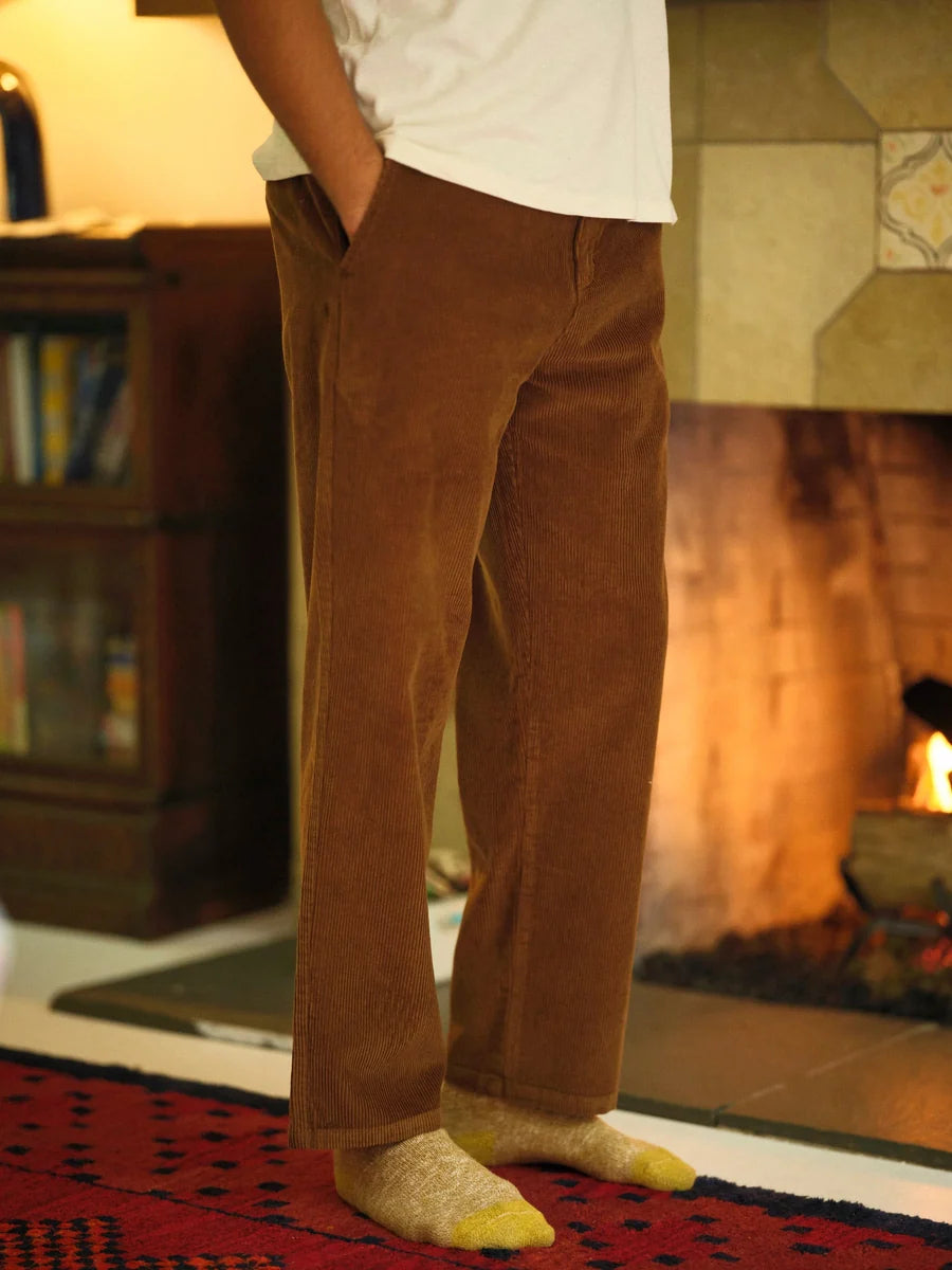 Front view from the waist down of a man wearing the Mollusk Surf Shop Boat Pants in Brown Wide Wale Corduroy, showing the relaxed straight-leg fit, structured front with zip fly, side hand pocket, and rich brown medium-wale corduroy fabric, styled with a white tee and yellow socks in front of a lit fireplace
