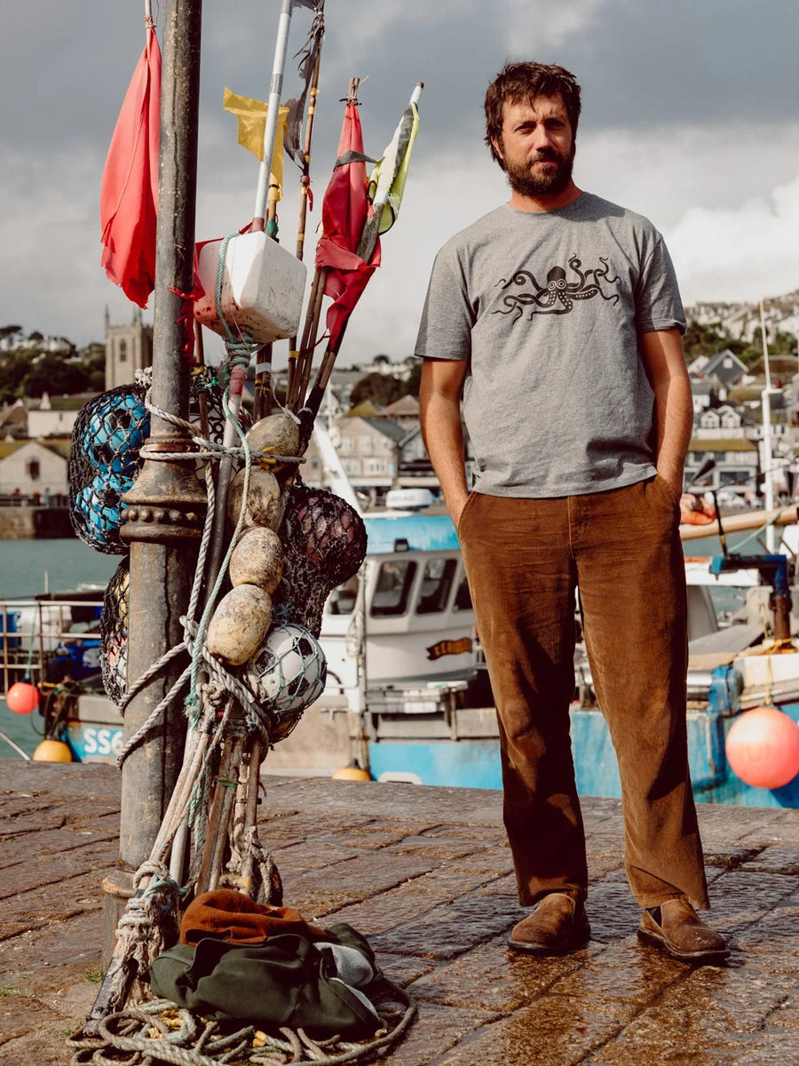Full-length lifestyle shot of a man wearing the Mollusk Boat Pants in Brown Wide Wale Corduroy with a gray graphic tee and brown suede shoes, standing on a harbor dock beside a post hung with fishing buoys and colorful flags, with fishing boats and a coastal town in the background
