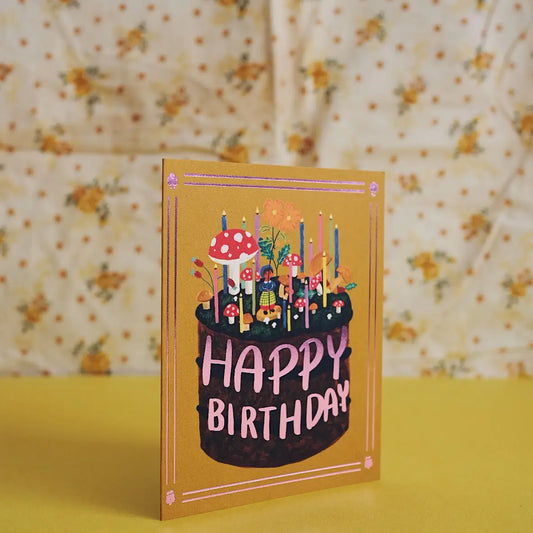Small Adventure Mushroom Cake Birthday Card, illustrated cake topped with mushrooms and colorful candles, with “Happy Birthday” lettering on a mustard-yellow background.
