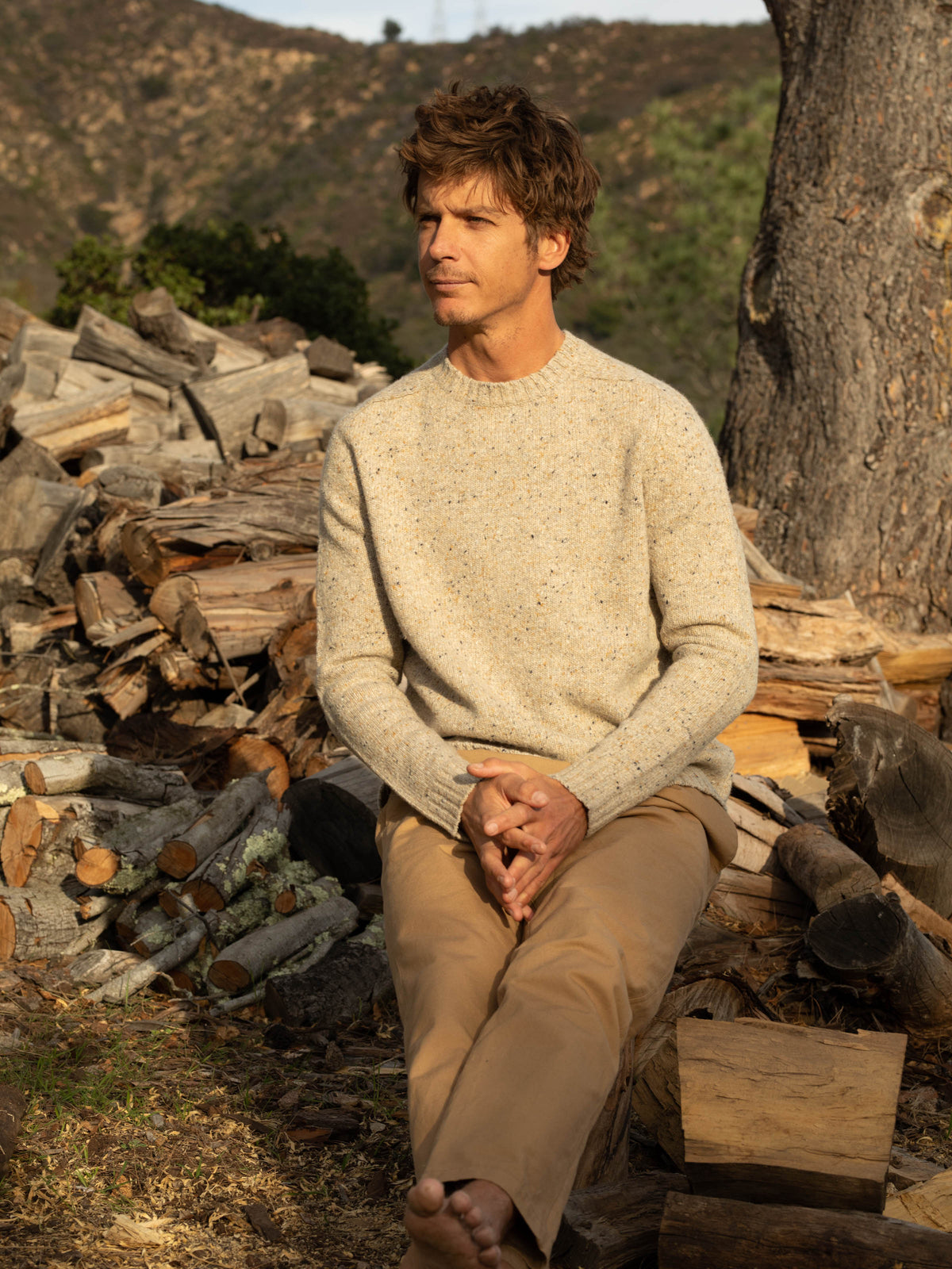 Full-length front view of a man seated barefoot on a log outdoors wearing the Mollusk Cambridge Sweater in Oat, showing the slim crew neck fit, saddle shoulder, and Donegal oat knit with multicolor fleck, styled with tan chino pants beside a stacked firewood pile and a large tree trunk at golden hour
