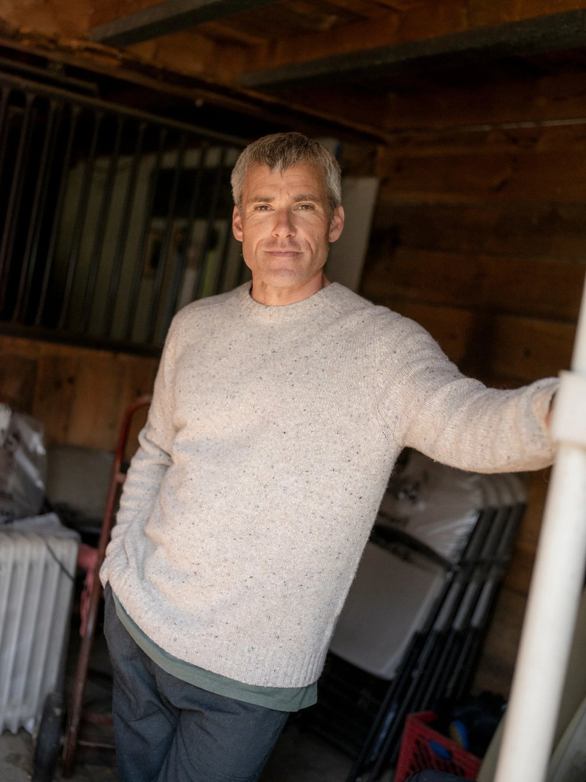 Front portrait of a man leaning against a post in a rustic barn interior wearing the Mollusk Cambridge Sweater in Oat, showing the slim crew neck fit and Donegal oat knit with multicolor fleck, styled with dark gray pants with a gray layer visible at the hem
