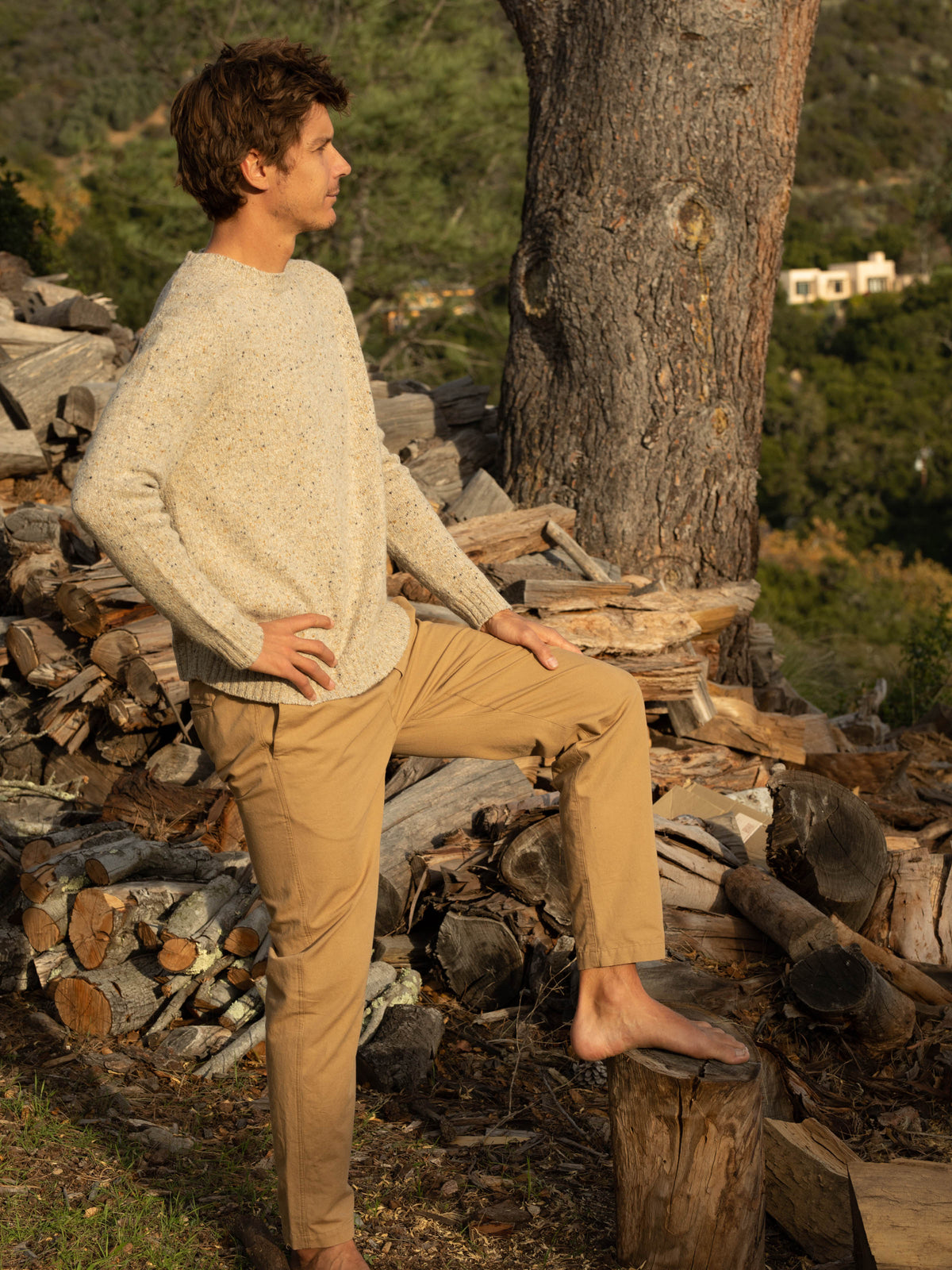 Full-length side view of a man standing barefoot on a log pile outdoors wearing the Mollusk Cambridge Sweater in Oat, showing the slim silhouette, ribbed cuffs and hem, and Donegal oat knit, styled with tan chino pants against a hillside landscape at golden hour
