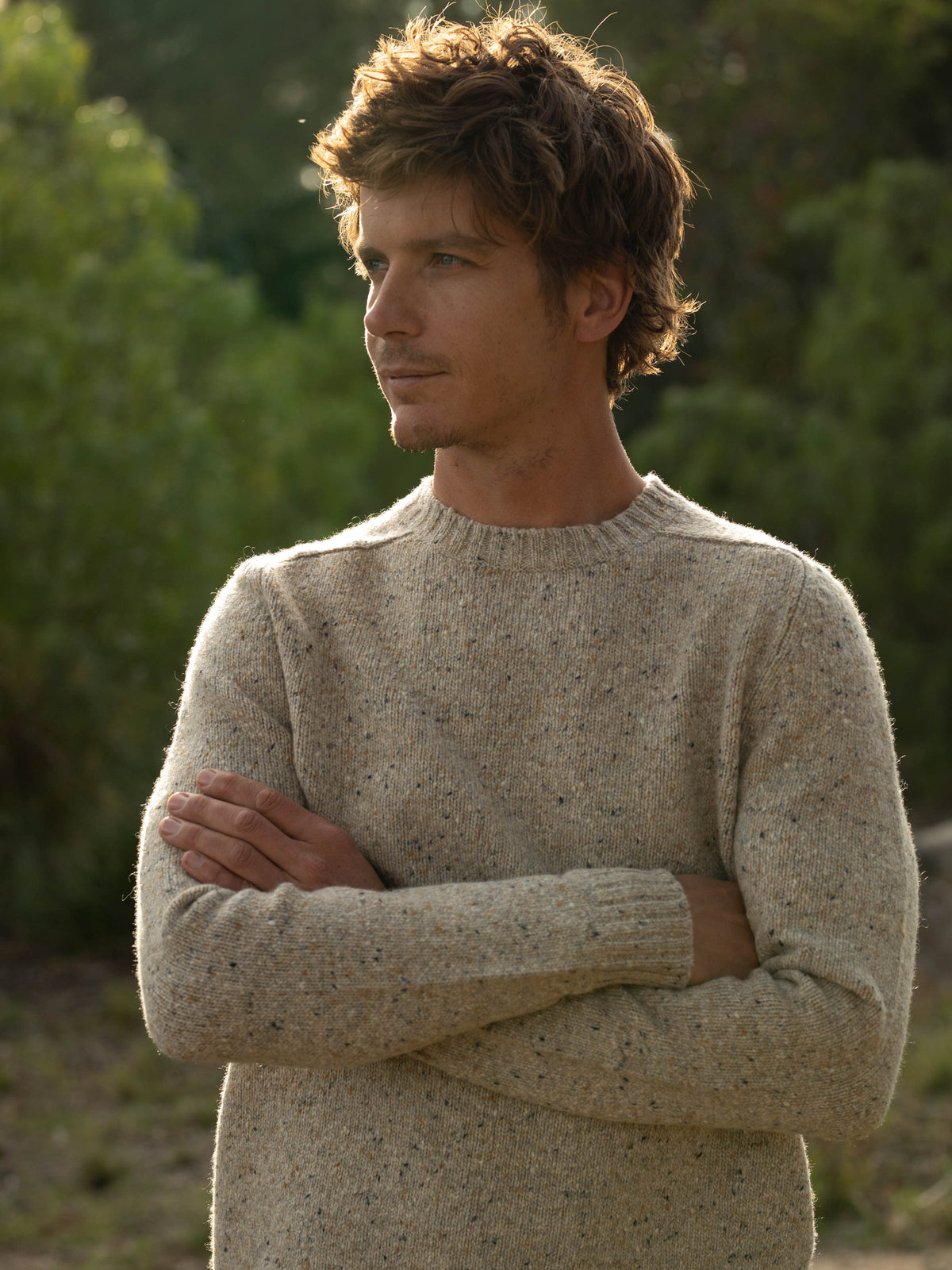 Portrait of a man standing outdoors with arms crossed wearing the Mollusk Surf Shop Cambridge Sweater in Oat, showing the slim-fitting crew neck, saddle shoulder seam, and Donegal knit in warm oat with multicolor fleck, against a soft-focus green hillside at golden hour
