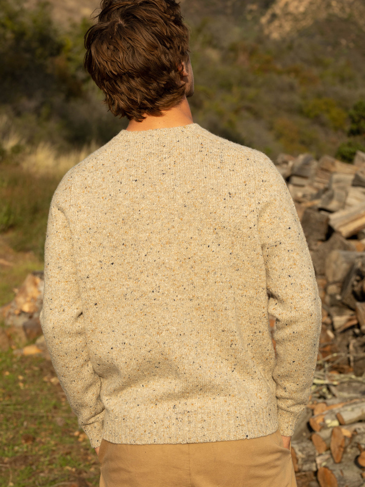 Back view of a man standing outdoors wearing the Mollusk Cambridge Sweater in Oat, showing the saddle shoulder construction, ribbed crew neck, and Donegal knit texture in warm oat with multicolor fleck, with a rocky hillside and stacked firewood in the background
