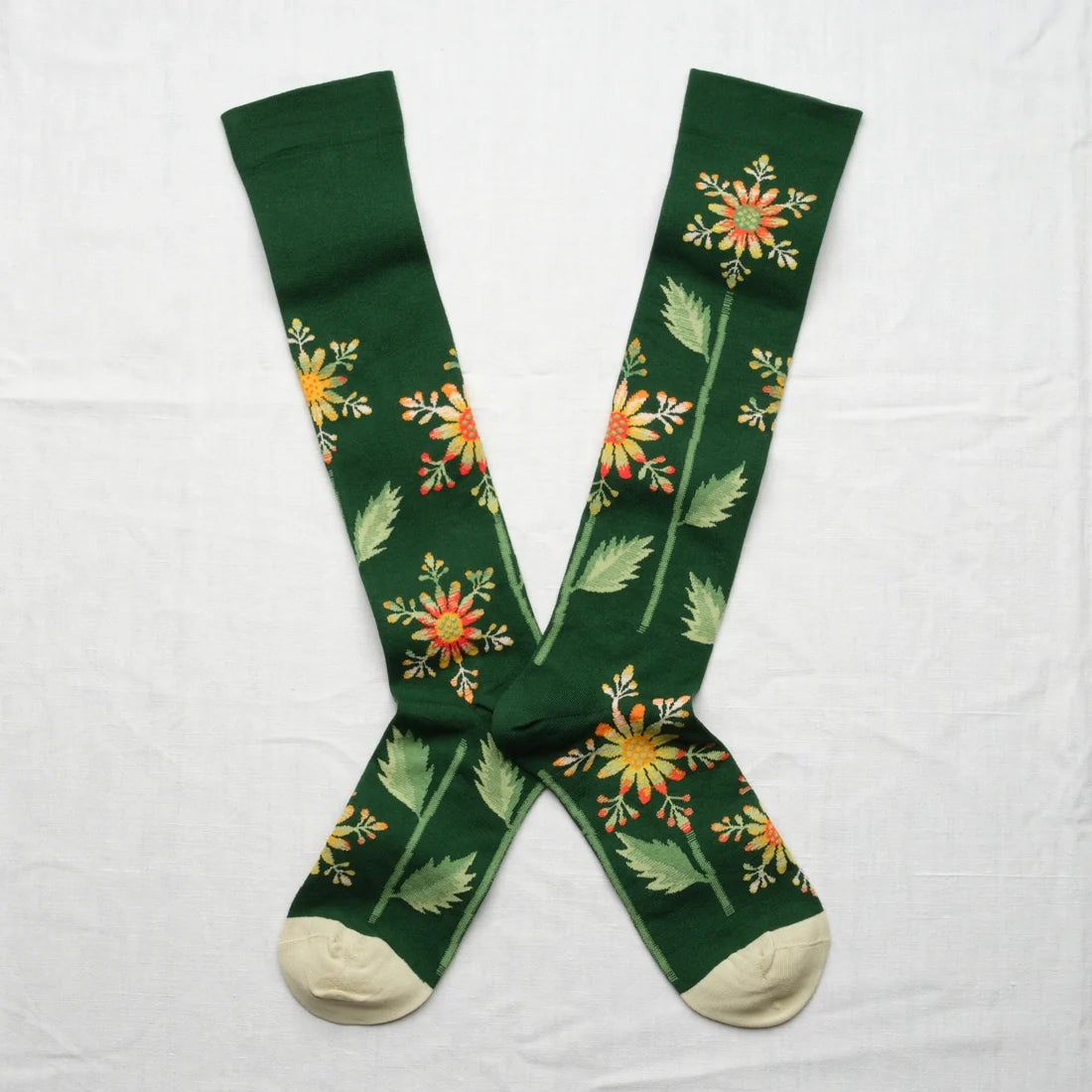 Bonne Maison Forest Snowflake Knee High Socks, flat lay pair showing forest green body with scattered snowflake-like botanical flower print in yellow, orange, and red with cream heel and toe
