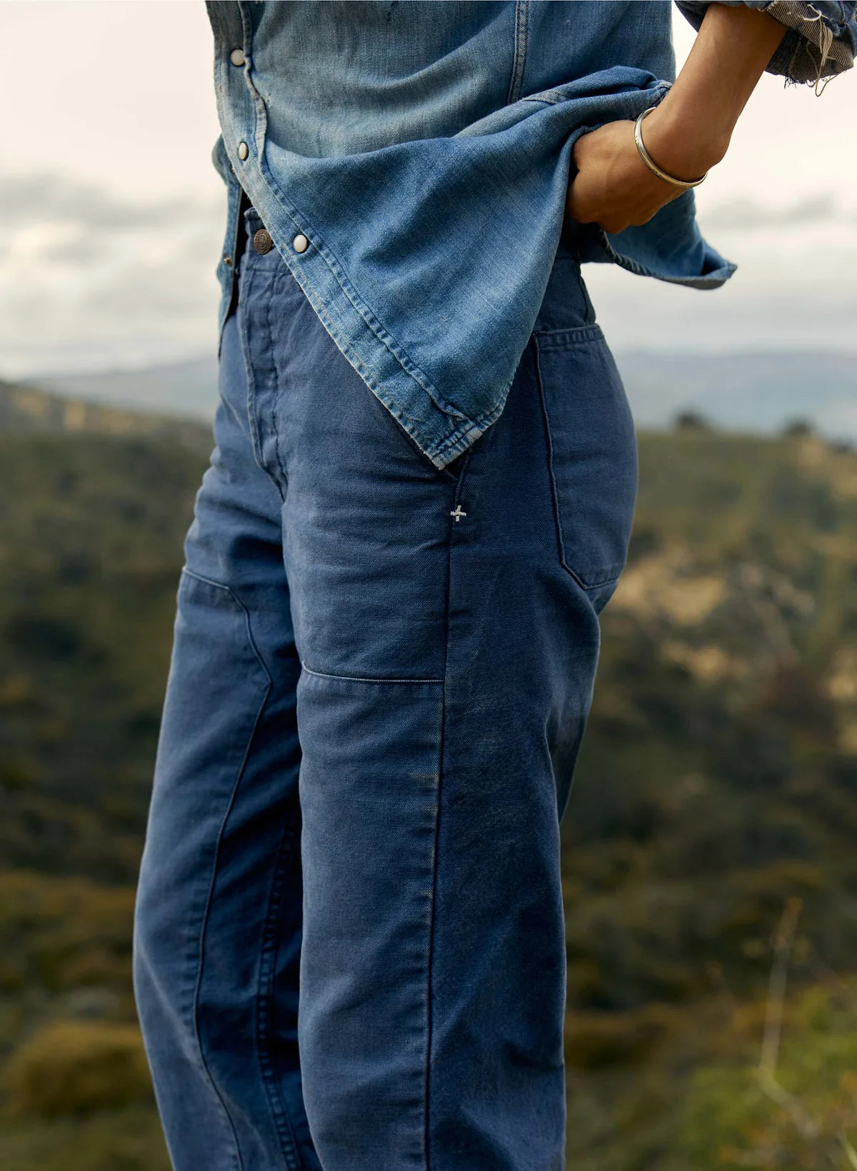 Close-up of blue cotton canvas workpant showing high-rise waist, double-knee panel, and patch pocket detail, styled with an open chambray shirt outdoors.
