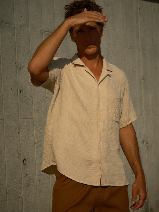 Man shading his eyes from the sun, wearing a oat colored shirt and brown pants against a concrete wall.