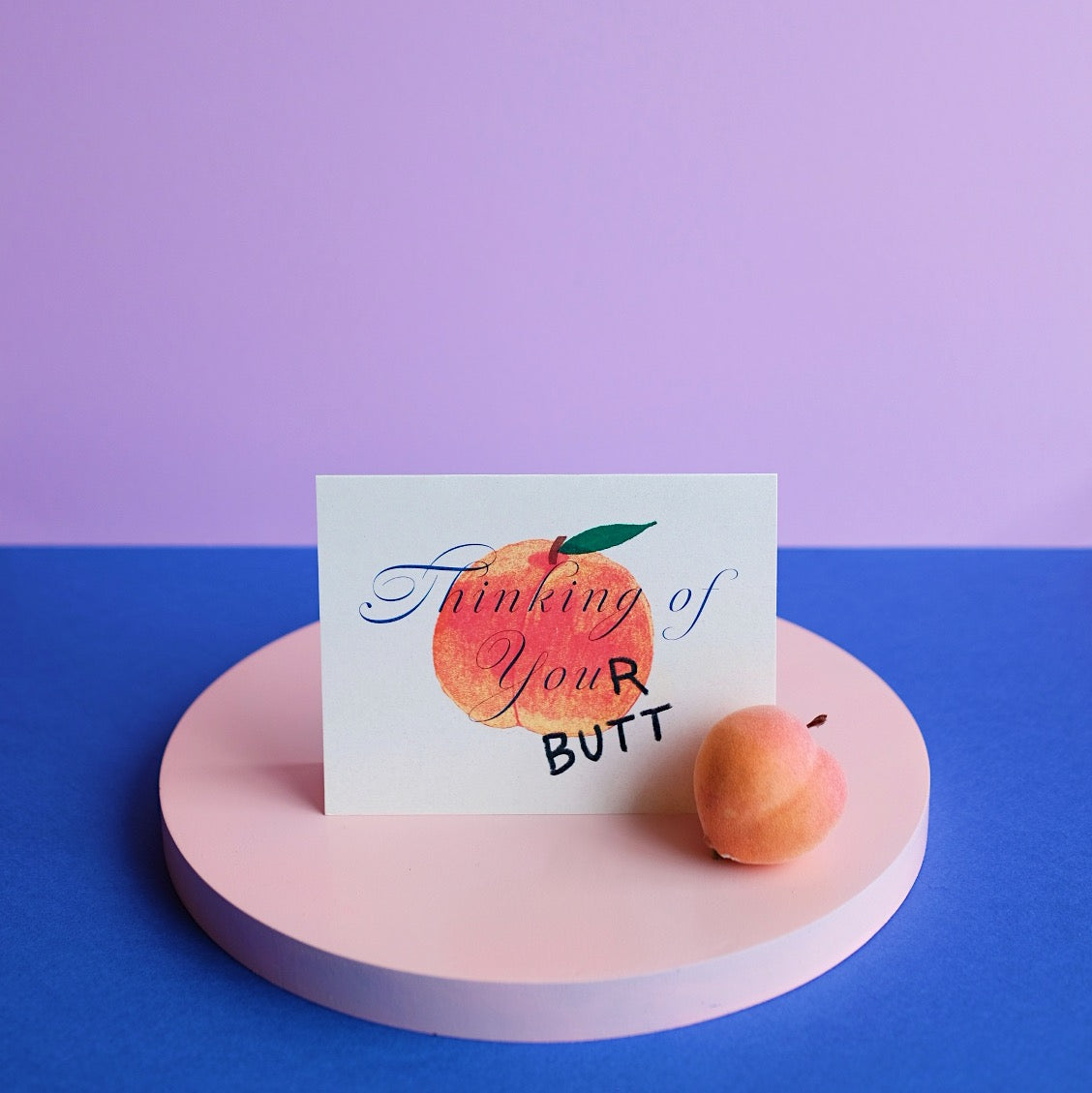 Small Adventure Thinking of Your Butt Card, peach illustration and “Thinking of Your Butt” lettering, displayed beside a peach on a pink stand.

