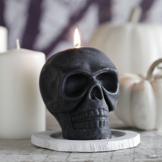 Big Dipper Wax Works Black Skull Beeswax Candle lit on a round white wood slice, front three-quarter view showing detailed anatomical skull shape in matte black with a burning flame at the crown, styled with white pillar candles and a small white pumpkin on a grey surface against a purple and white bokeh background
