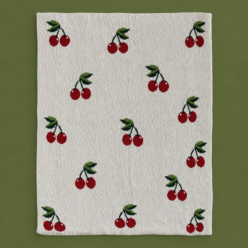 Flat lay of the Dotter Cotton Knit Baby Blanket with Cherries, showing the cream knit face with an allover intarsia pattern of red cherries with green stems and leaves, on a sage green background

