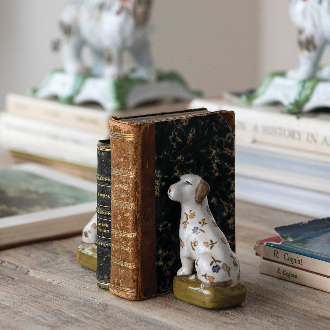 A ceramic dog bookend figurine with a white body and gold floral pattern sits on a green base, holding a vintage brown book and a black marbled book on a wooden table, with other dog figurines and books blurred in the background.