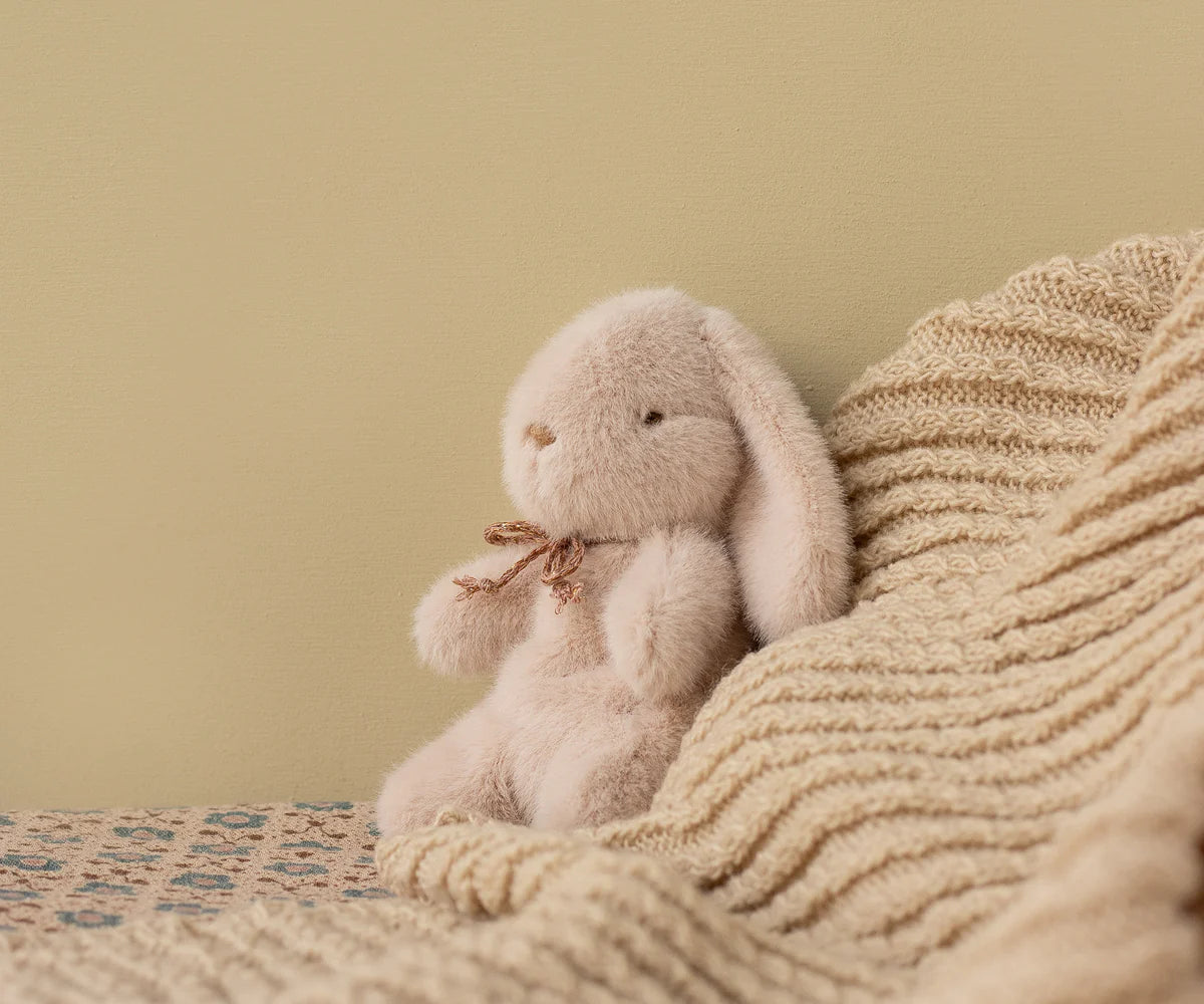 Plush bunny toy with a bow tie sitting on a textured blanket against a beige wall.