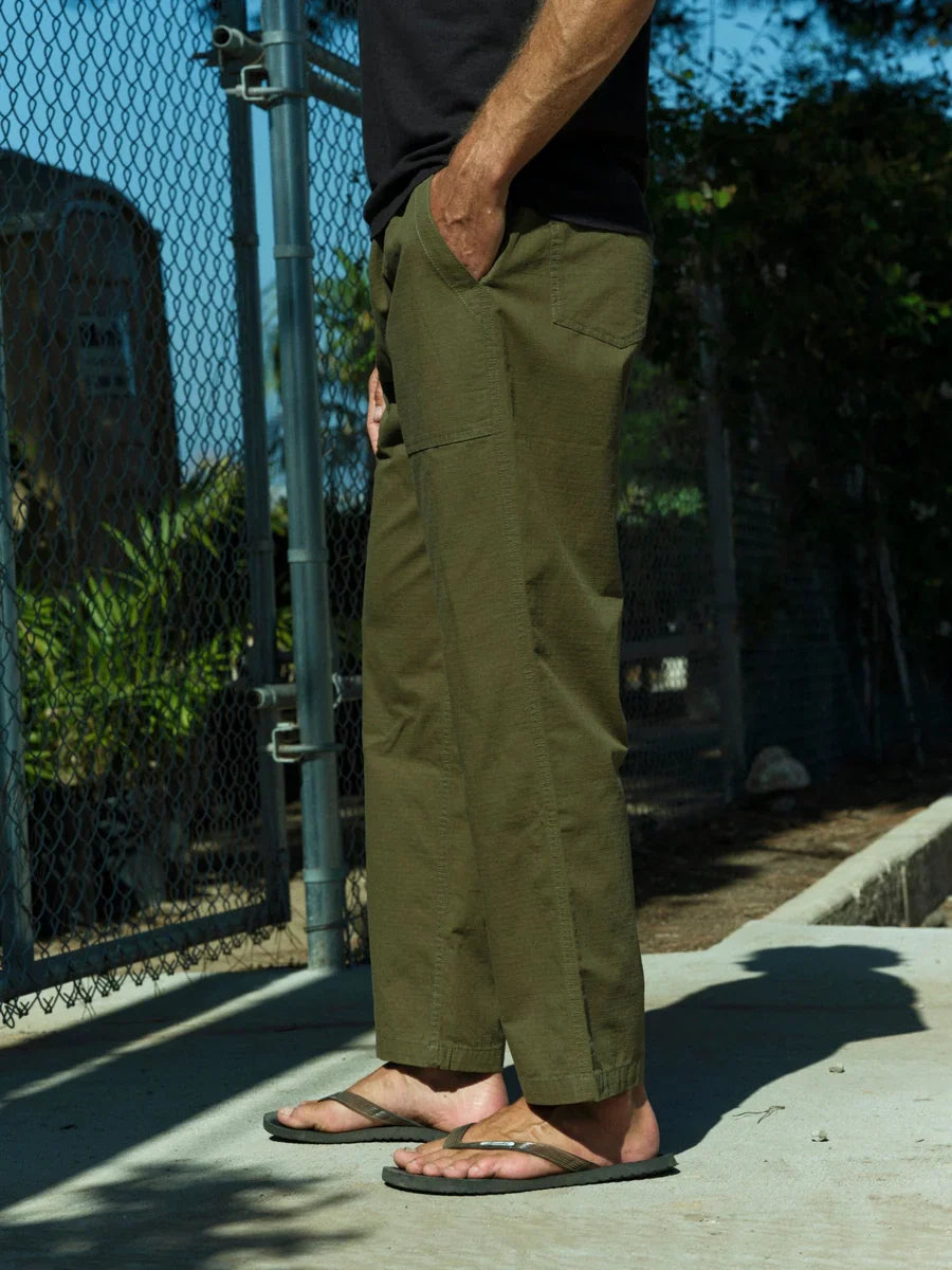 Model wearing Mollusk Surf Shop Drawstring Salvador Pants in Mash Green Ripstop, side view with hand in front slash pocket showing olive green cotton ripstop, roomy straight-leg silhouette, side vent at hem, and relaxed full-length drape, styled with black t-shirt and olive flip flops outdoors
