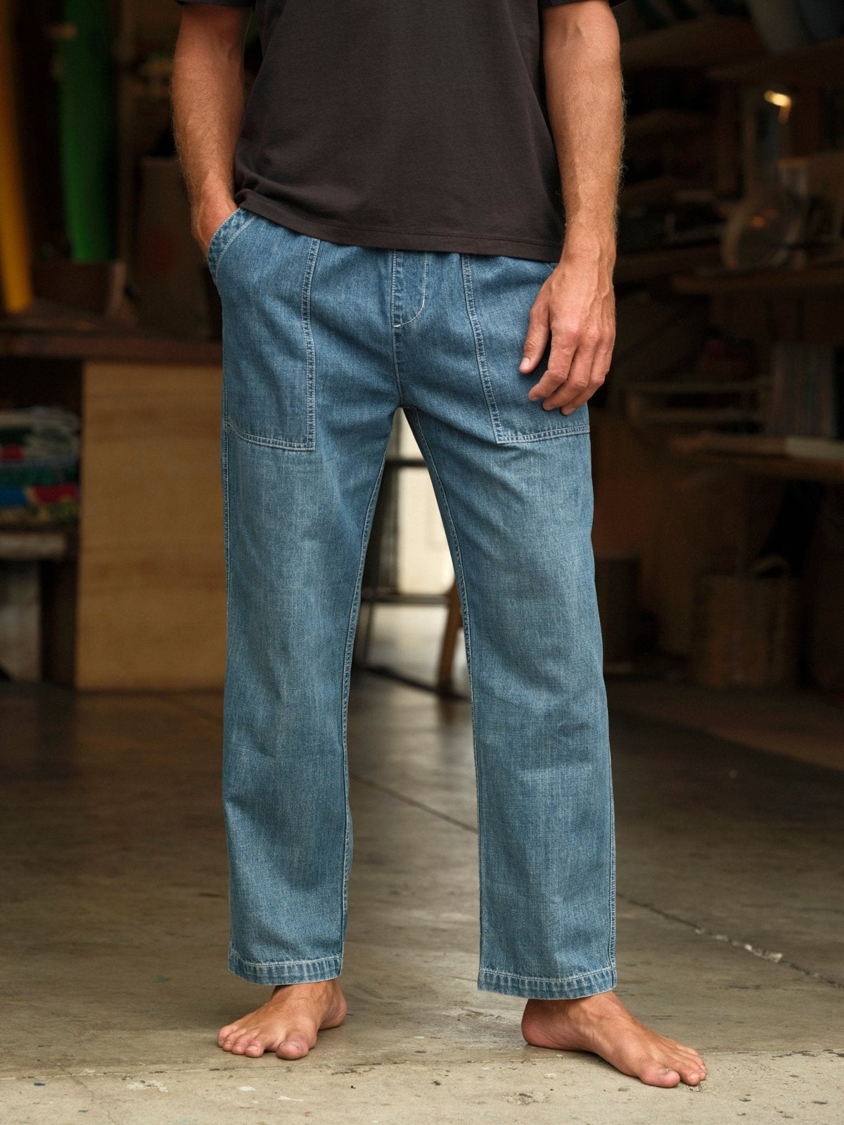 Model wearing Mollusk Surf Shop Drawstring Salvador Pants in Vintage Indigo Denim, front cropped view showing vintage indigo lightweight cotton denim, roomy straight-leg silhouette, elastic drawstring waistband, front patch pockets with carpenter panel seam detail on thigh, barefoot in a surf shaping room
