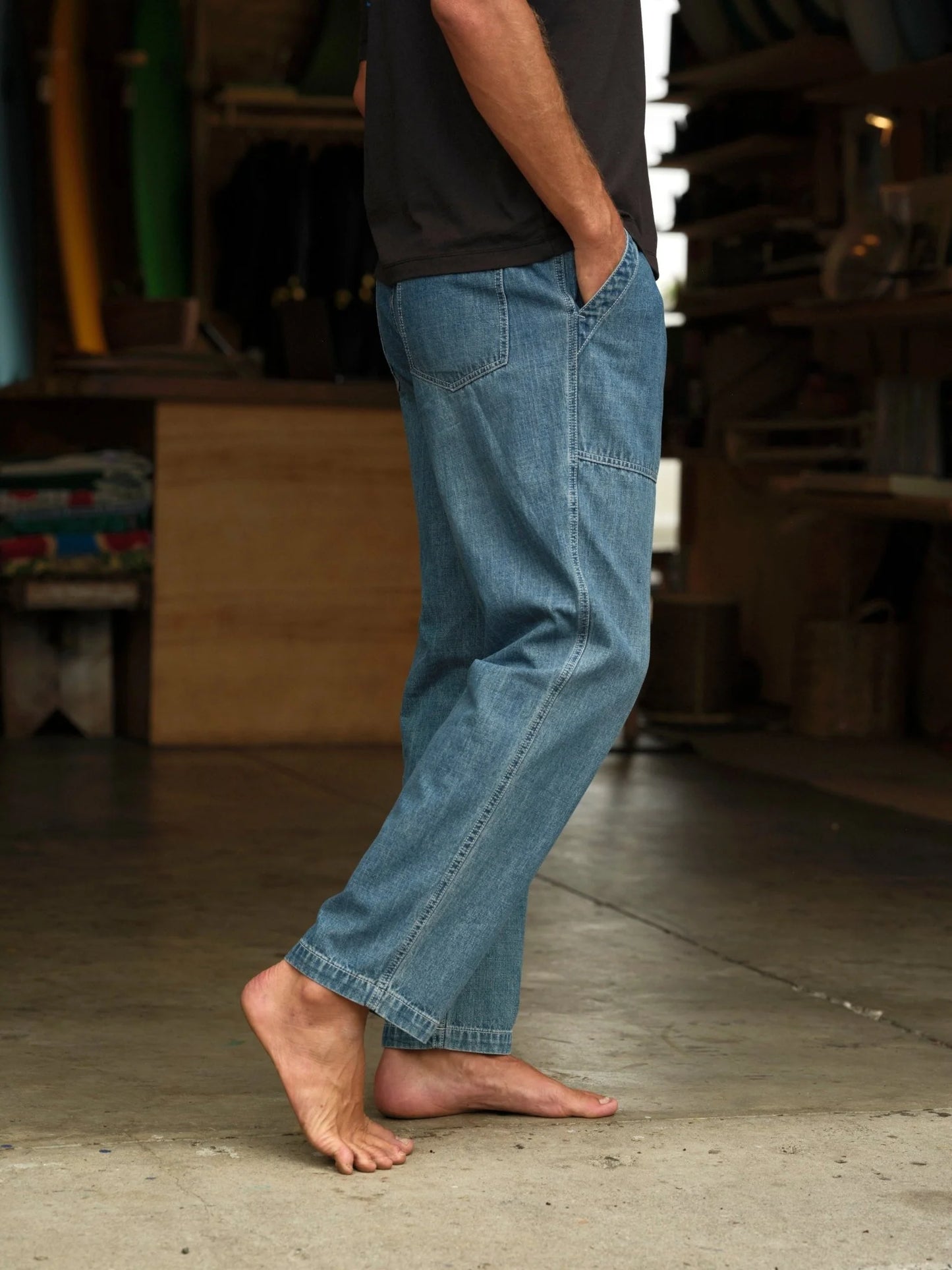 Model wearing Mollusk Surf Shop Drawstring Salvador Pants in Vintage Indigo Denim, side view walking barefoot in surf shaping room showing vintage indigo lightweight cotton denim, roomy straight-leg silhouette, side patch pocket, carpenter panel seam on thigh, and straight full-length hem, styled with dark t-shirt
