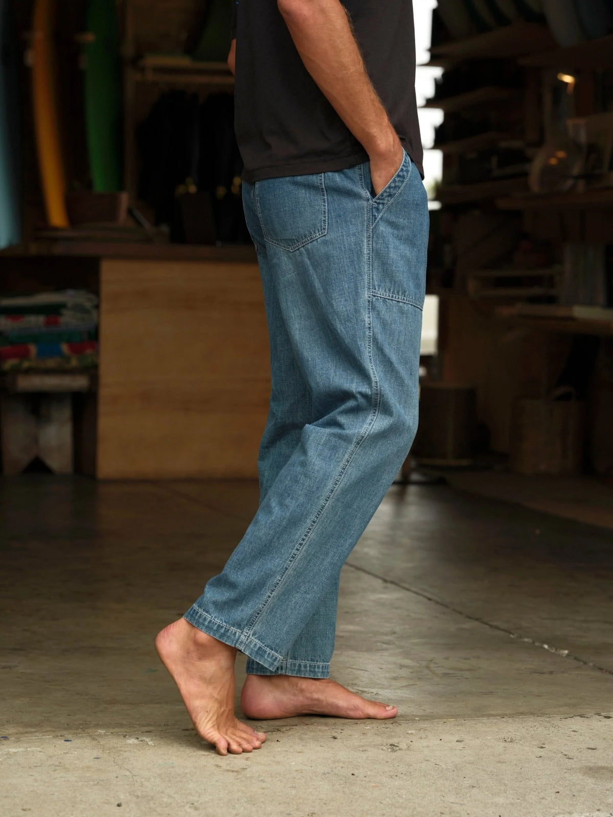 Model wearing Mollusk Surf Shop Drawstring Salvador Pants in Vintage Indigo Denim, side view walking barefoot in surf shaping room showing vintage indigo lightweight cotton denim, roomy straight-leg silhouette, side patch pocket, carpenter panel seam on thigh, and straight full-length hem, styled with dark t-shirt
