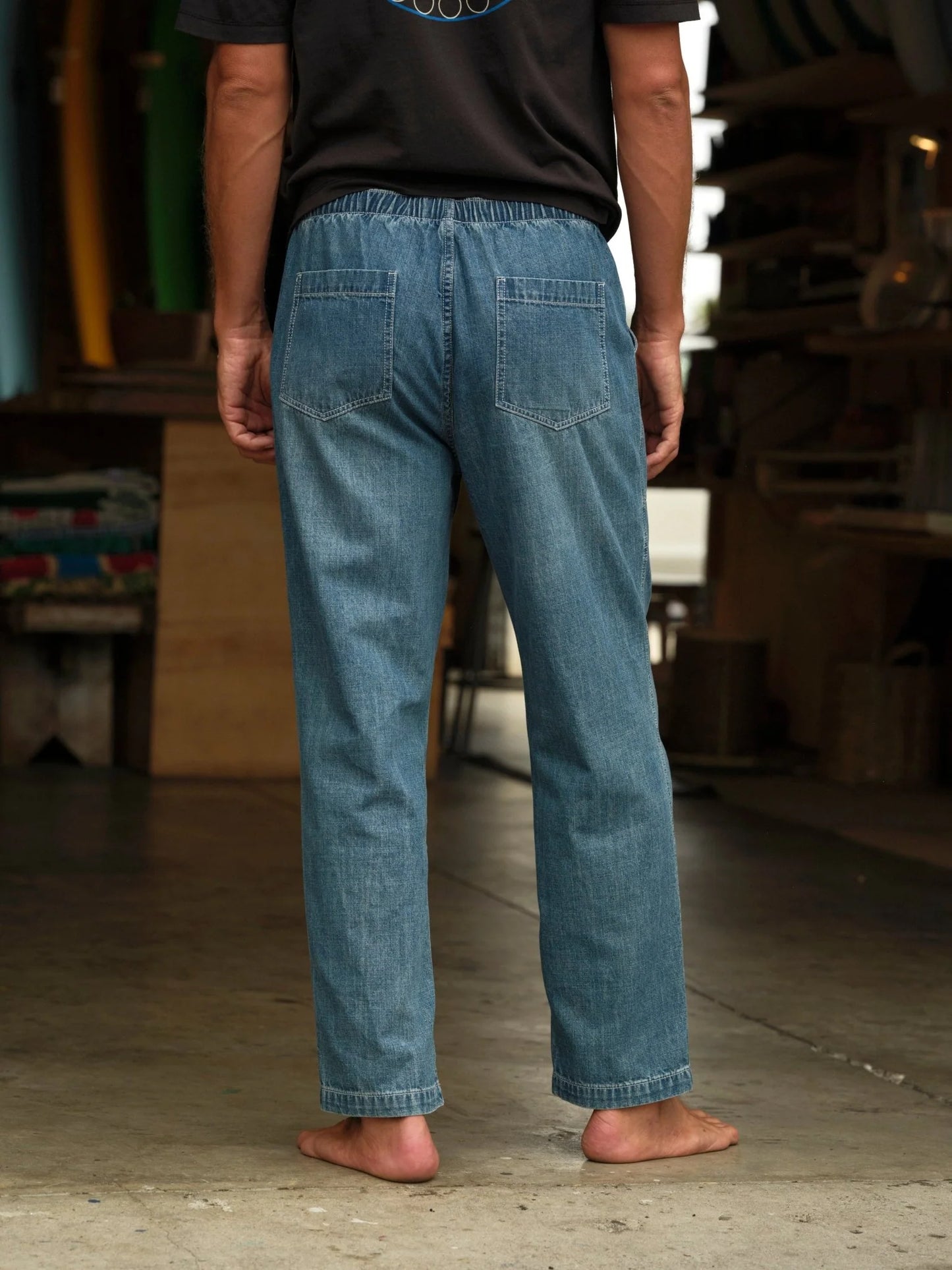 Model wearing Mollusk Surf Shop Drawstring Salvador Pants in Vintage Indigo Denim, back view barefoot in surf shaping room showing elastic drawstring waistband, two back patch pockets, roomy straight-leg silhouette, and vintage indigo lightweight denim with straight full-length hem, styled with dark Mollusk graphic t-shirt
