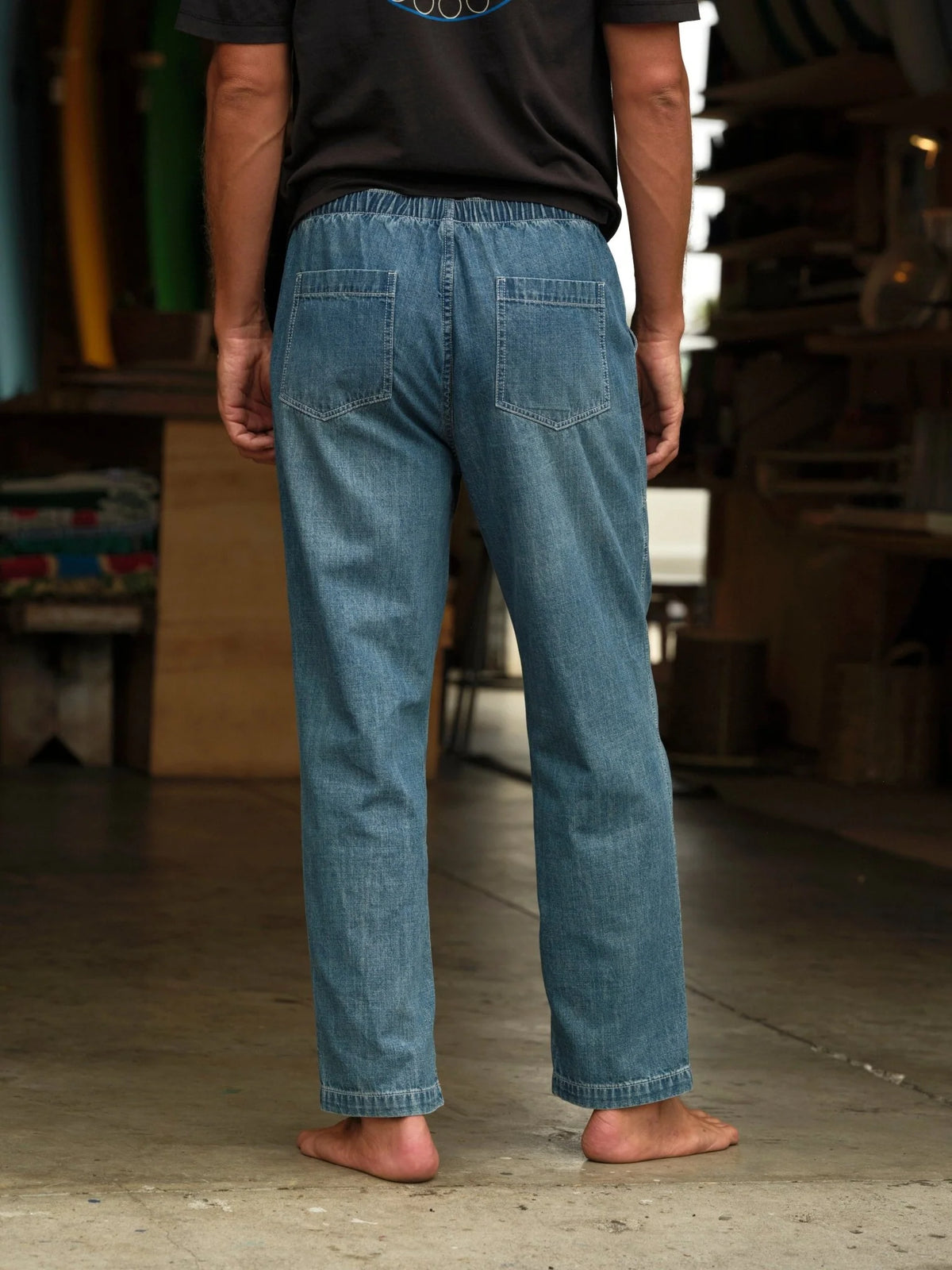 Model wearing Mollusk Surf Shop Drawstring Salvador Pants in Vintage Indigo Denim, back view barefoot in surf shaping room showing elastic drawstring waistband, two back patch pockets, roomy straight-leg silhouette, and vintage indigo lightweight denim with straight full-length hem, styled with dark Mollusk graphic t-shirt
