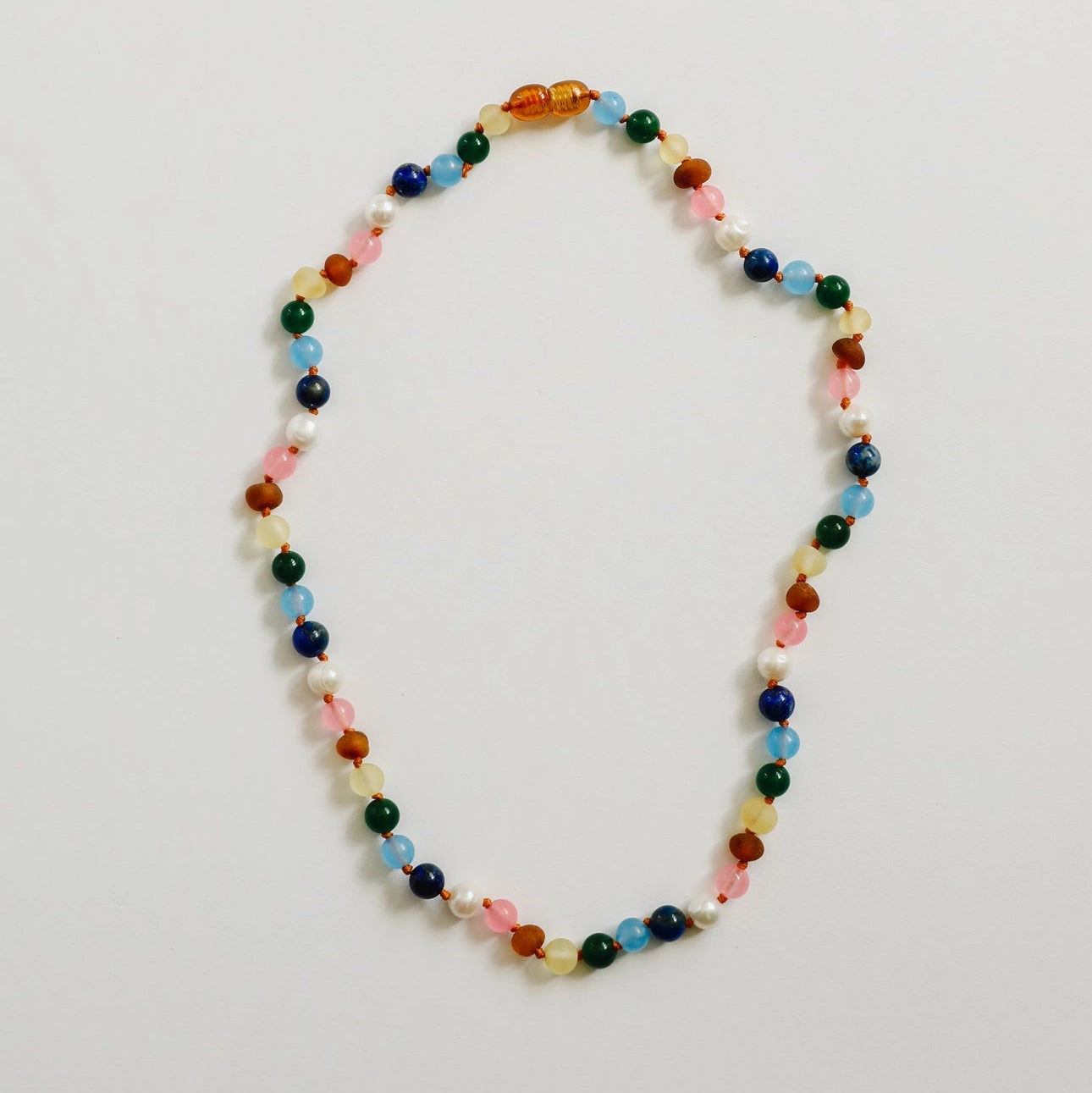 CanyonLeaf Raw Baltic Amber and Pearl + Multi Colored Gemstones Necklace — a hand-knotted necklace alternating raw honey amber, white pearl, pink rose quartz, blue aquamarine, navy lapis, green jade, and yellow amber beads with a honey amber screw clasp, laid flat on a light grey background