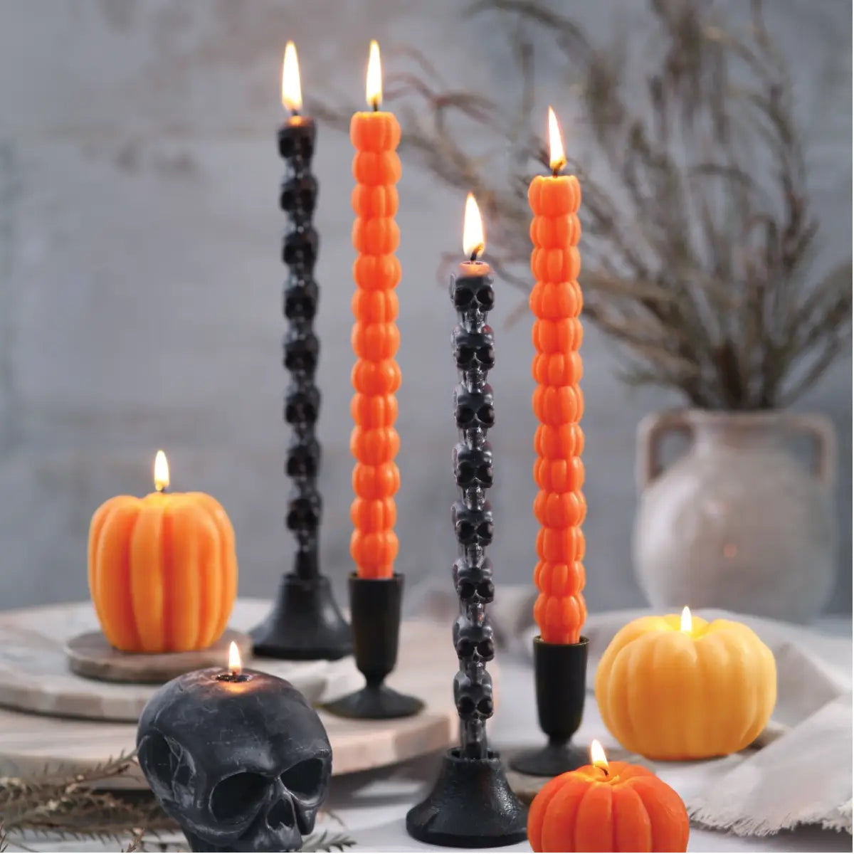 Big Dipper Wax Works Sculpted Black Skull Taper Candles lit and displayed in black candlestick holders on a Halloween tablescape alongside orange pumpkin-shaped candles, dried branches in a ceramic vase, and a black skull prop, against a moody grey wall background
