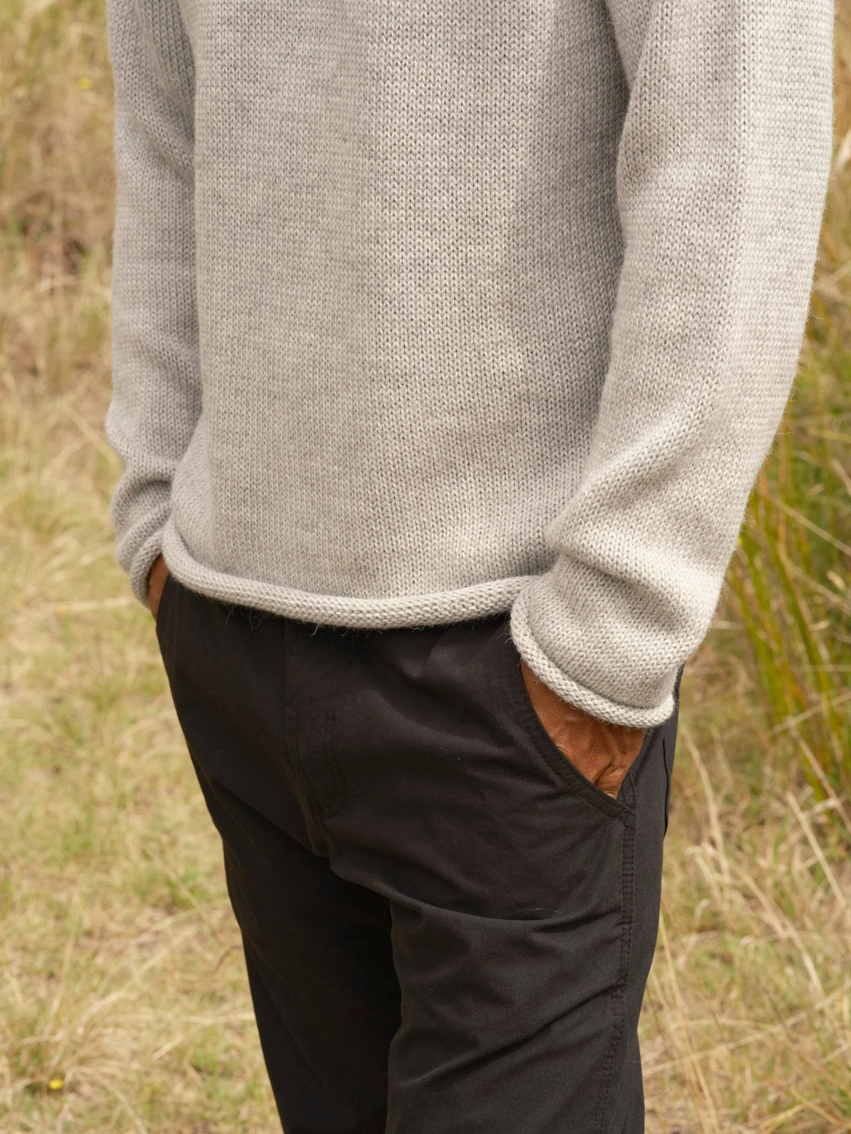 Detail shot of the lower body and hem of a man wearing the Mollusk Fisherman Sweater in Pearl Gray, showing the rolled hem finish, heathered light gray alpaca blend knit texture, and rolled cuff, styled with black pants against a dry coastal meadow
