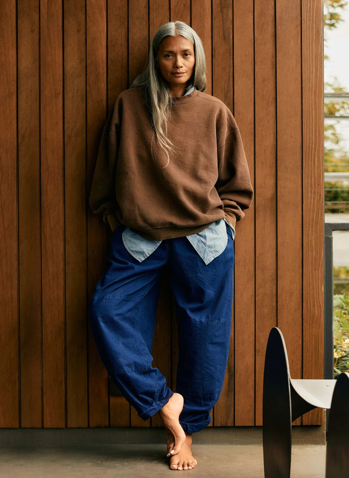 Model wearing indigo sashiko wide-leg flight pants with a brown oversized sweatshirt, leaning against a wood-paneled wall.
