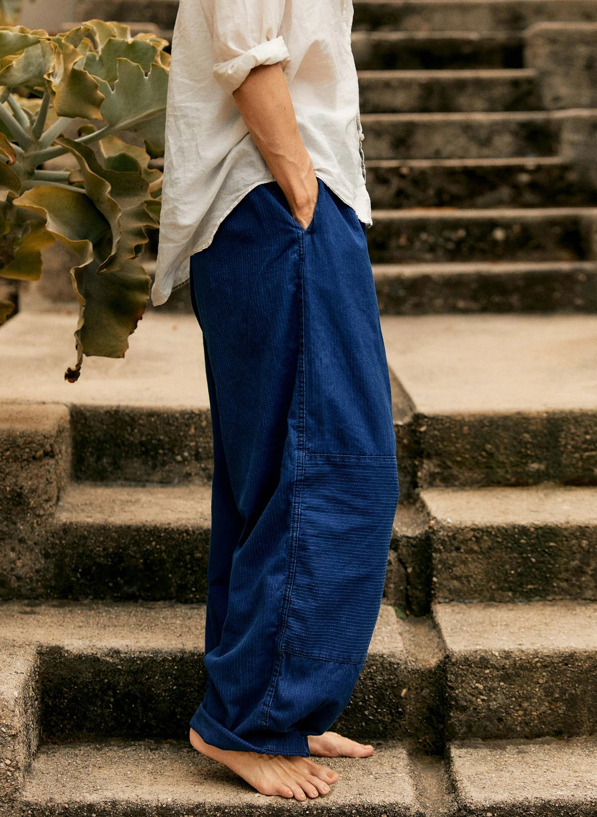 Side view of indigo sashiko flight pants showing the wide leg, reinforced double-knee panel, and side pocket, styled with a white linen shirt.
