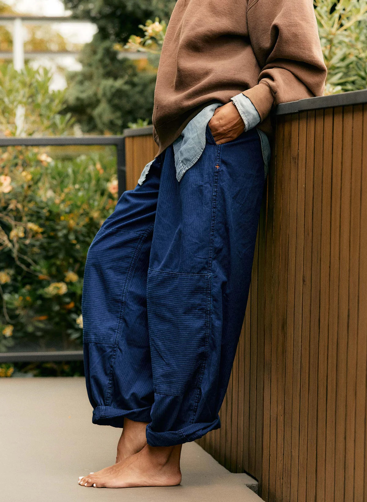 Close-up of the lower half of indigo sashiko flight pants showing the box-pleated hem and double-knee panel, styled with a brown sweatshirt outdoors.
