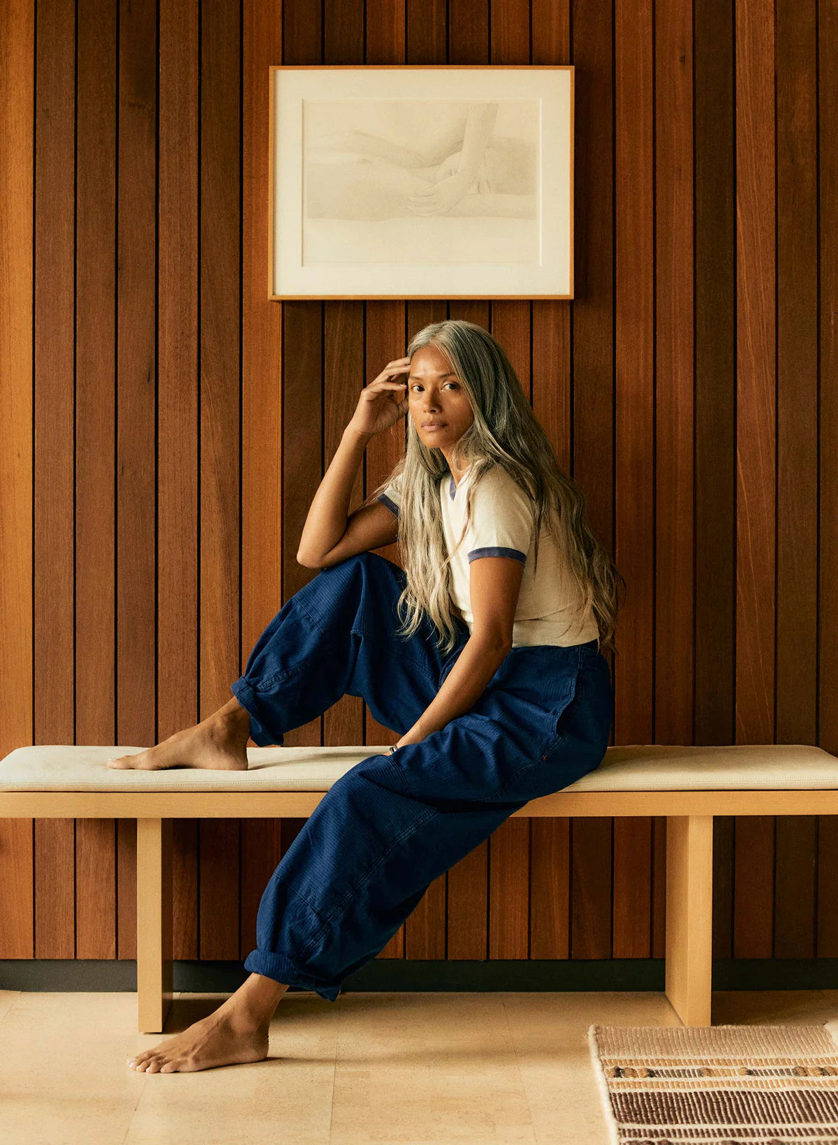Model seated on a wood bench wearing indigo sashiko flight pants and a cream ringer tee, against a warm wood-paneled wall.
