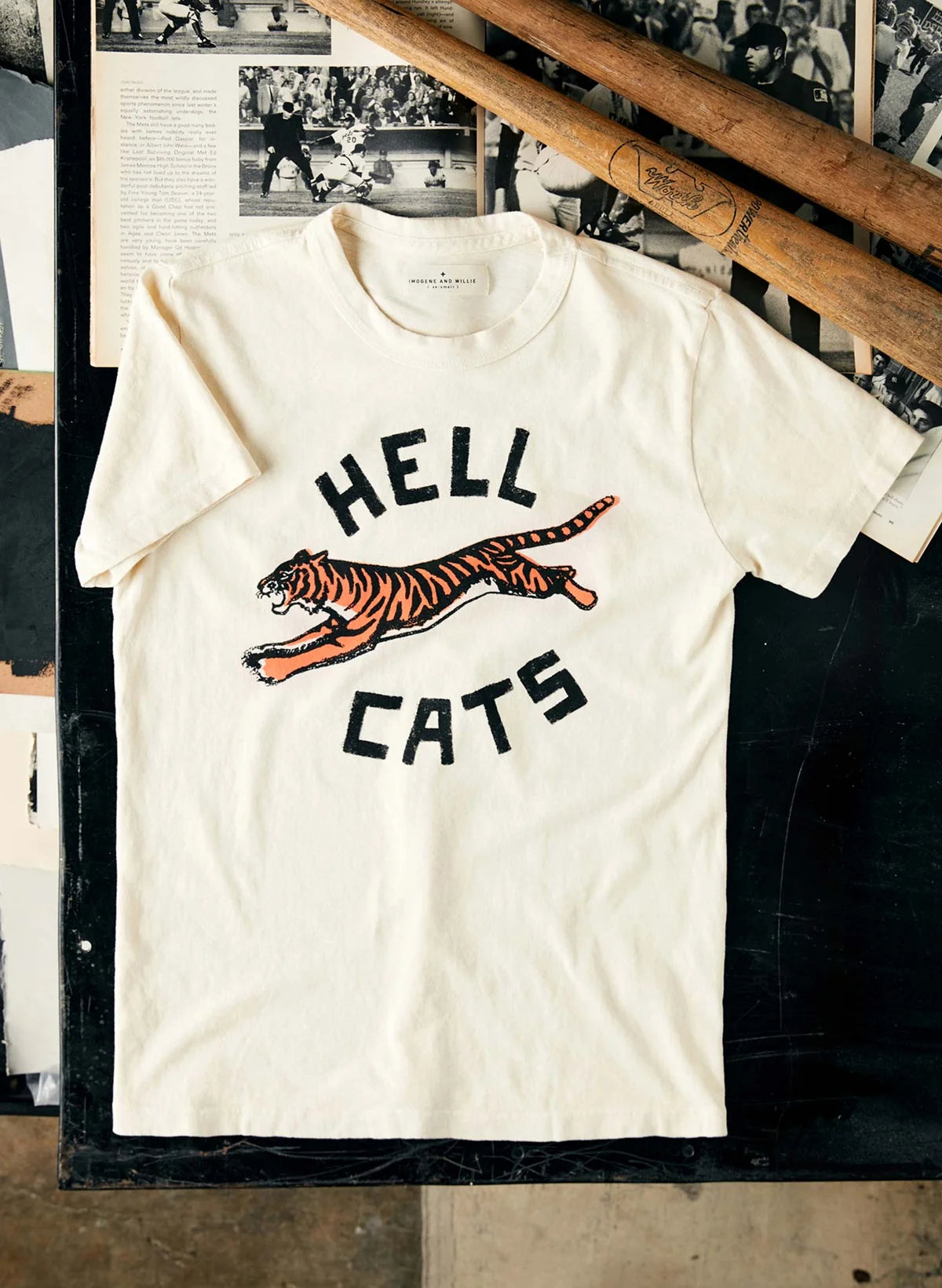 Cream Hellcats tee laid flat on a dark surface with a vintage baseball bat, featuring a leaping tiger screen print with "Hell Cats" text above and below.
