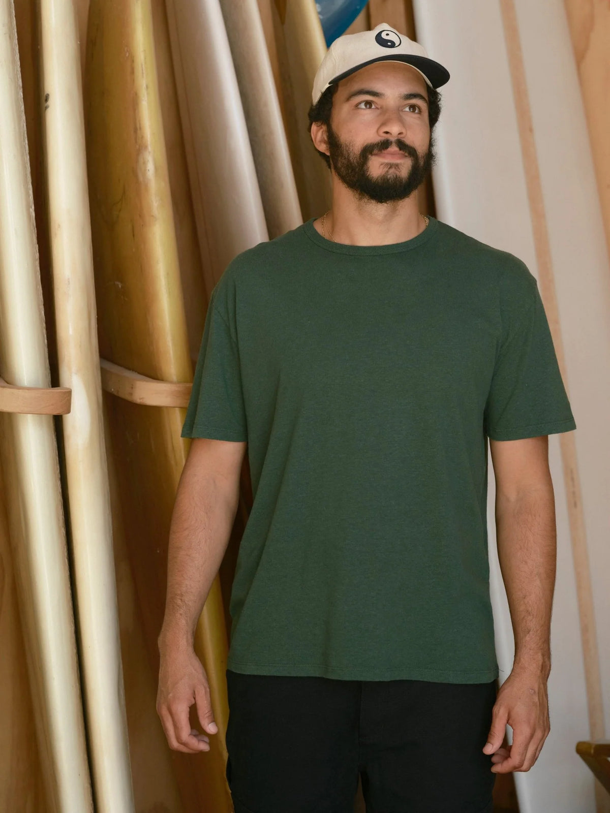 Front view of a man wearing the Mollusk Surf Shop Hemp Tee in Forest, showing a relaxed classic fit with ribbed crewneck and short sleeves in garment-dyed dark forest green 55% hemp 45% cotton jersey, styled with a yin-yang snapback cap in a wooden surfboard shaping room

