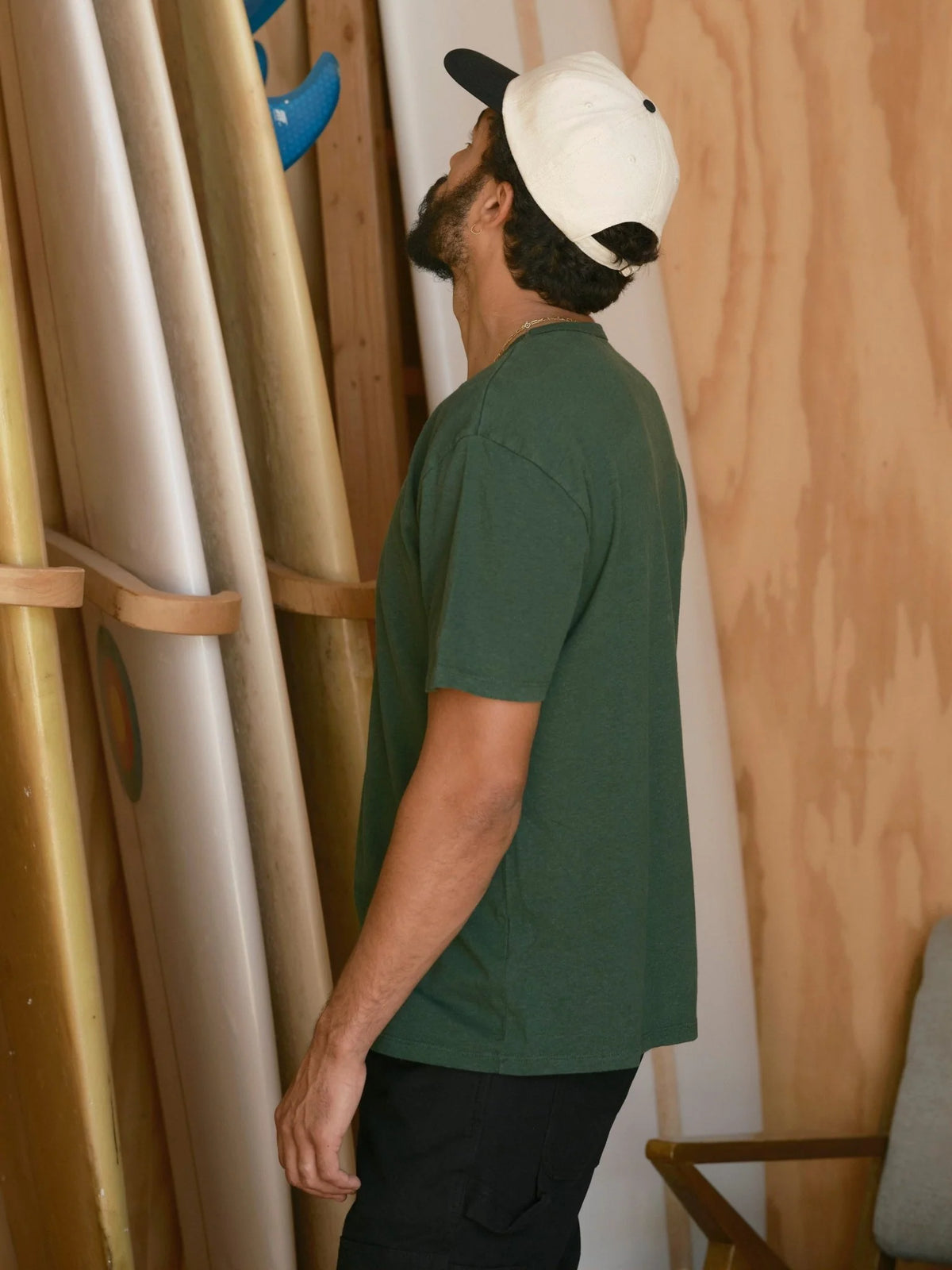 Back view of a man wearing the Mollusk Surf Shop Hemp Tee in Forest, showing the relaxed silhouette, short sleeves, and clean back seam in garment-dyed dark forest green hemp-cotton jersey, styled with a two-tone cap in a wooden surfboard shaping room

