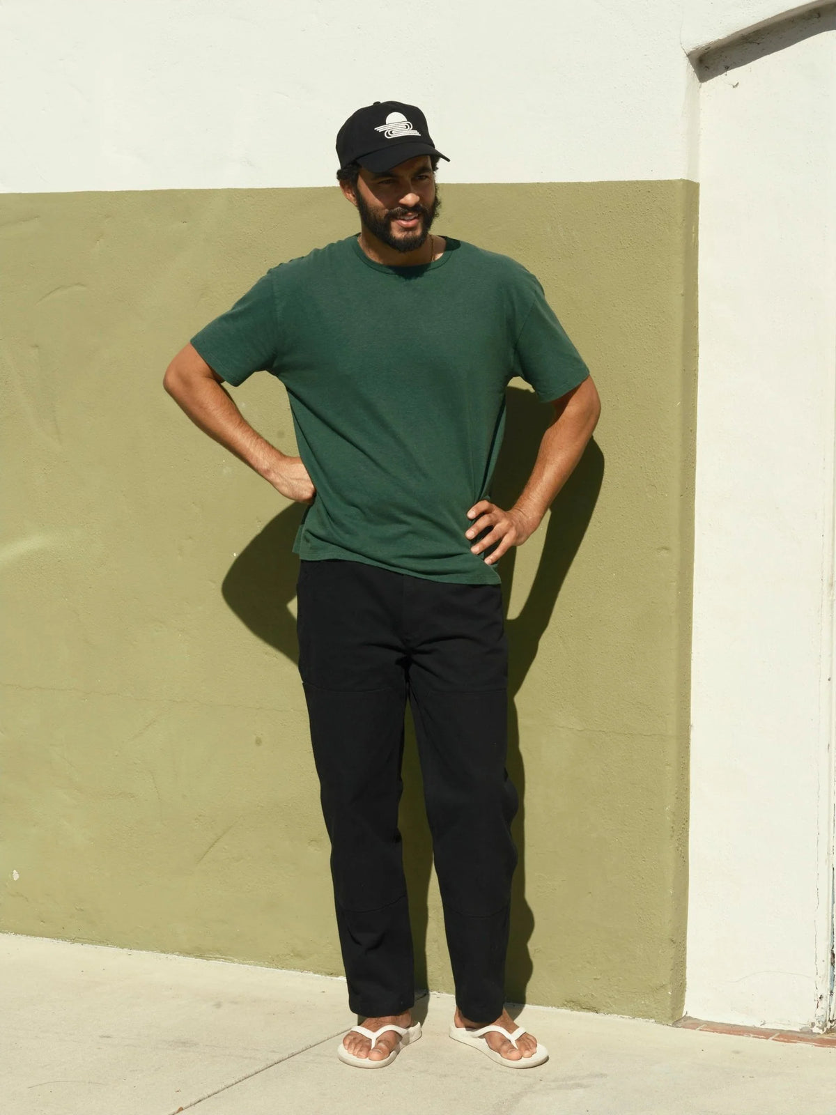 Full-length front view of a man wearing the Mollusk Hemp Tee in Forest with dark straight-leg pants and white sandals, standing with hands on hips against a khaki-painted outdoor wall in bright sunlight, showing the relaxed fit and garment-dyed color
