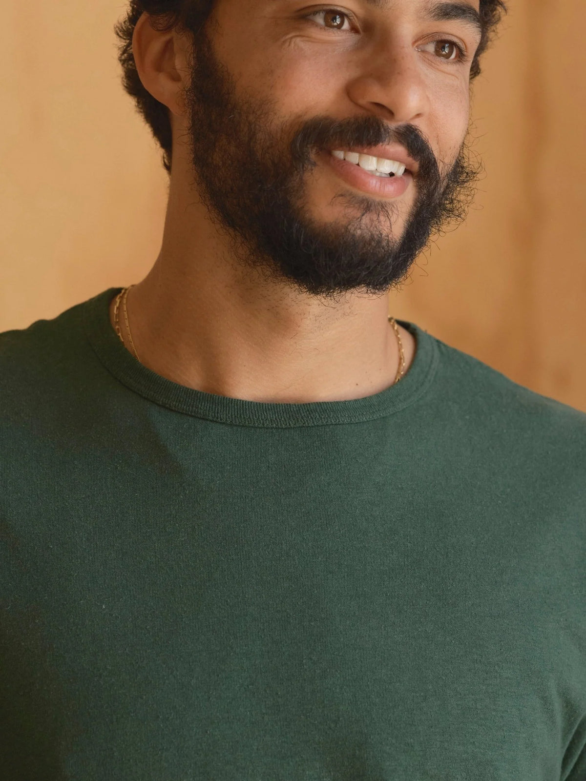 Close-up of the crewneck and chest of the Mollusk Hemp Tee in Forest, showing the ribbed neckline, fine jersey texture, and rich garment-dyed dark green color of the 55% hemp 45% cotton fabric against a warm wood-toned background
