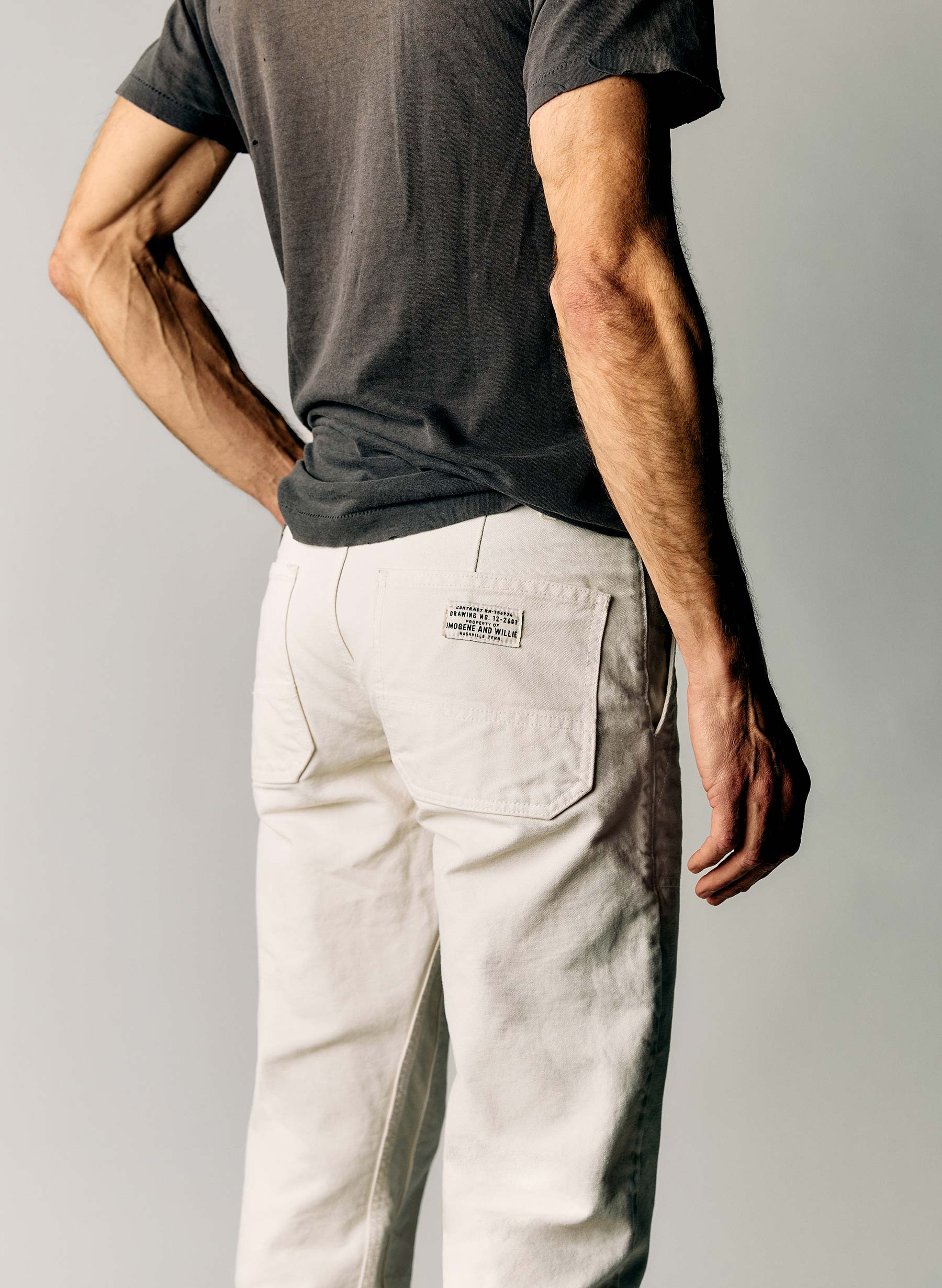 Back view close-up of the Hencye Canvas Workpant in Natural showing the large patch pocket with the Imogene and Willie label and the straight leg fit, worn with a charcoal tee