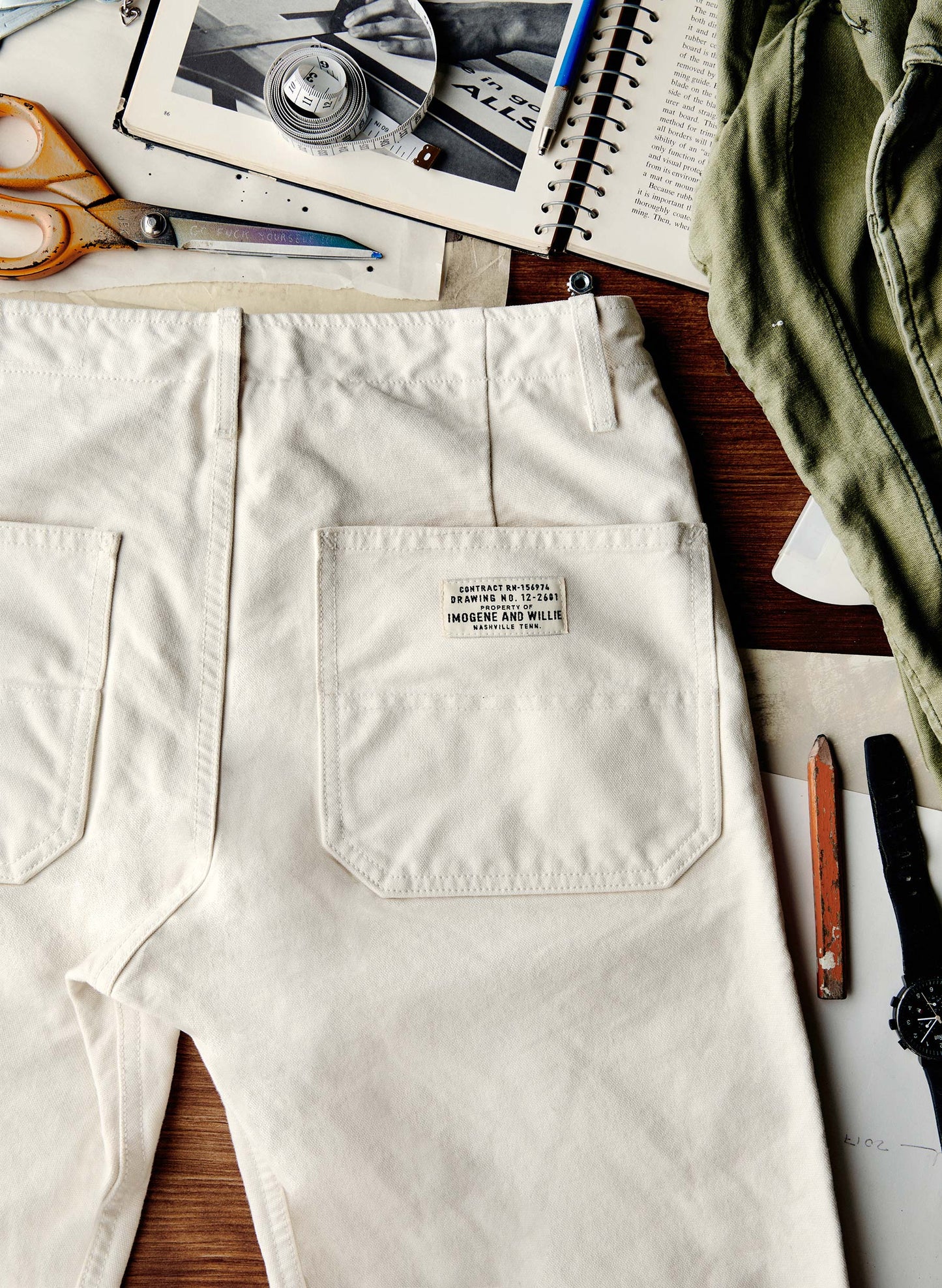 Close-up flat lay of the back of the Hencye Canvas Workpant in Natural showing the large patch pocket with the Imogene and Willie contract label, surrounded by workshop tools including scissors, a tape measure, and a pencil