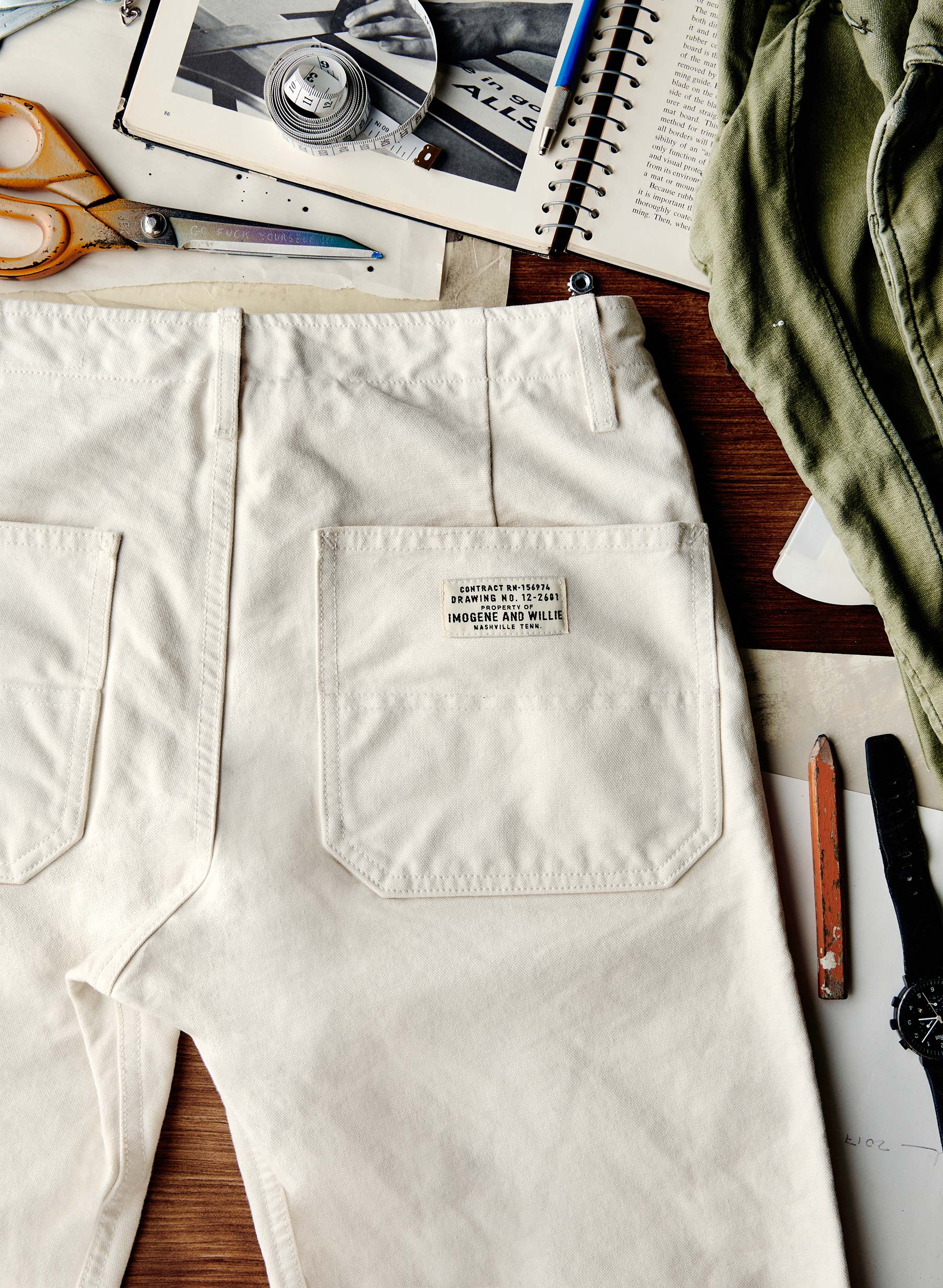 Close-up flat lay of the back of the Hencye Canvas Workpant in Natural showing the large patch pocket with the Imogene and Willie contract label, surrounded by workshop tools including scissors, a tape measure, and a pencil