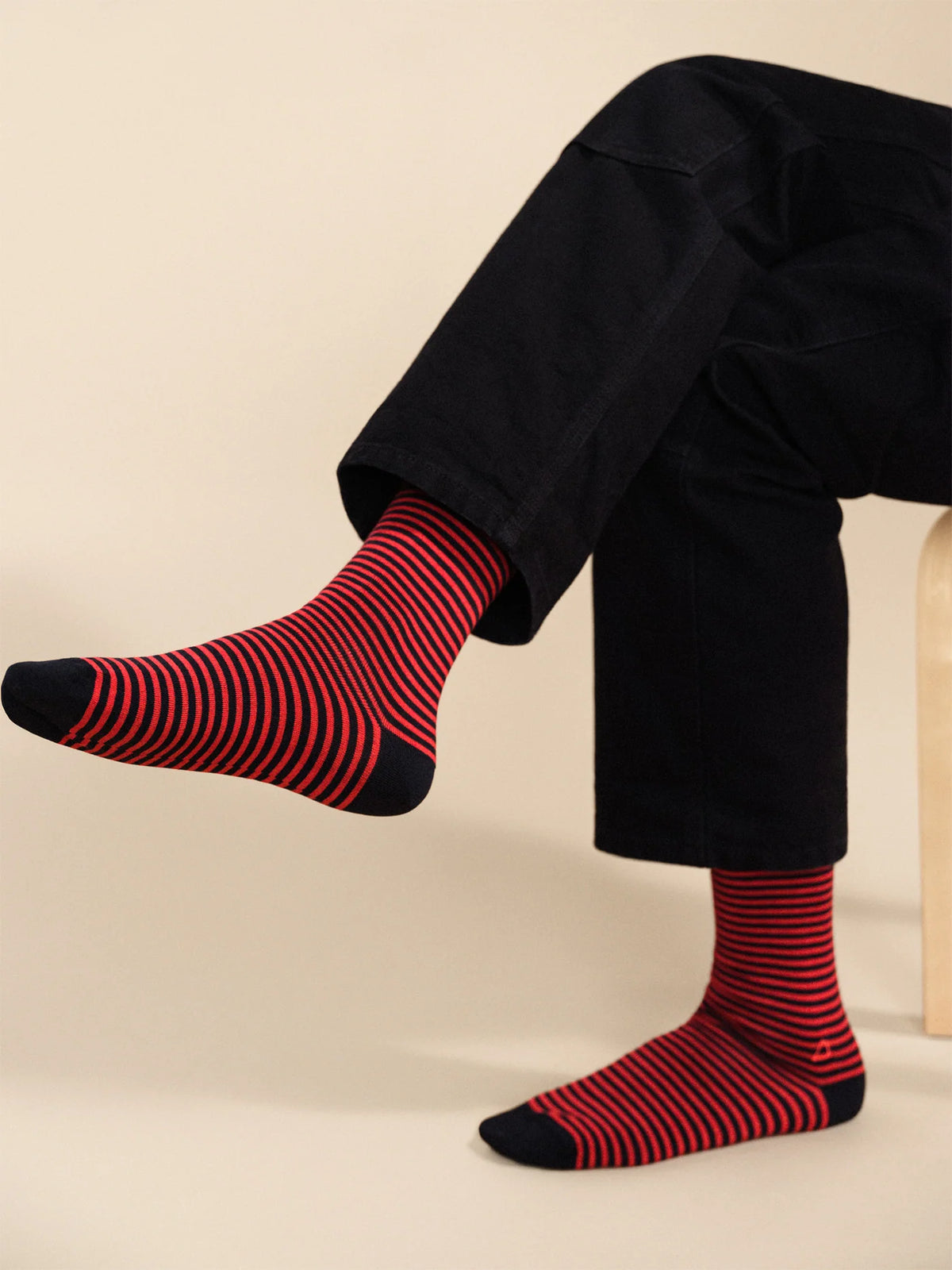 Model wearing navy and red striped crew socks with black trousers, seated with one foot raised.
