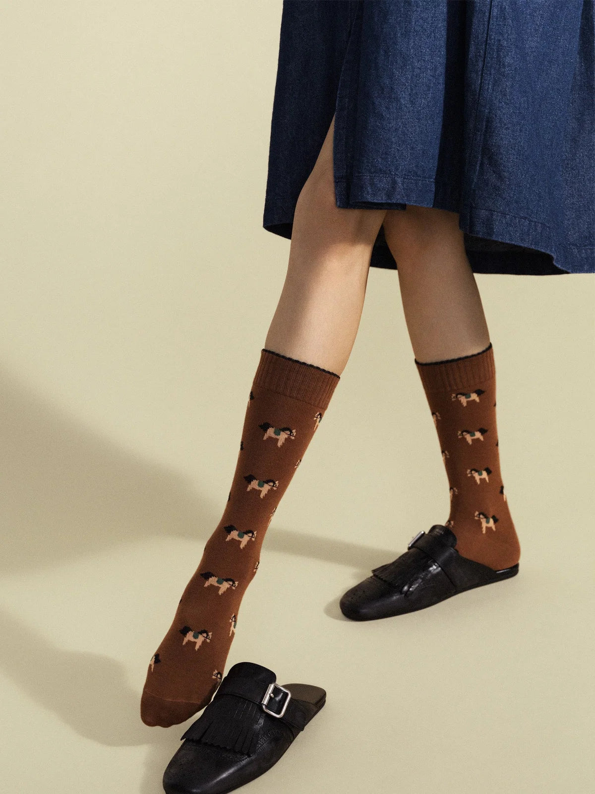 Equestrian Crew socks in Brown — rich brown cotton boot-height crew with allover equestrian horse motif, worn with black leather buckle mules under a navy denim midi skirt on a cream background
