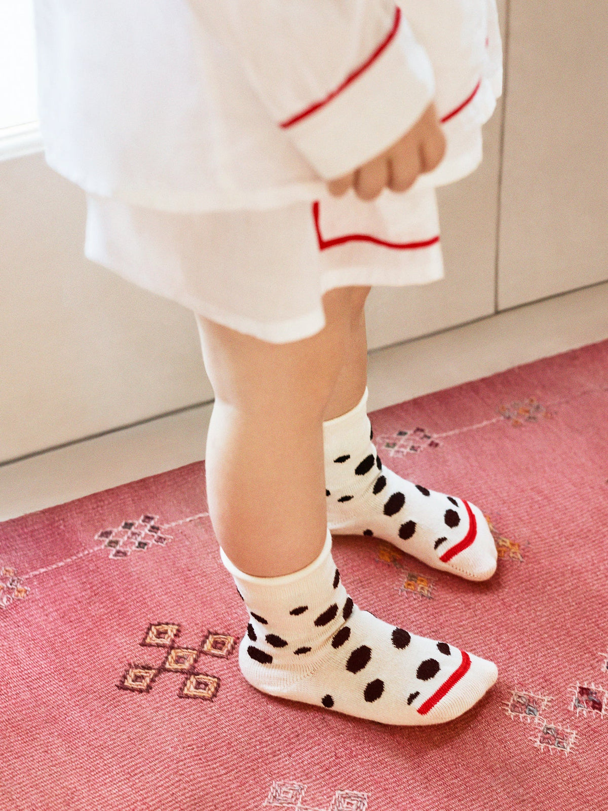 Child wearing ivory crew socks with black Dalmatian spots and red toe stripes, standing on a pink rug.
