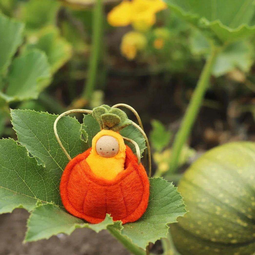 Fairy Shadow Pumpkin Seedling Necklace in light skin tone, resting on a large green pumpkin leaf in a garden, showing a knitted light-skin seedling doll in a yellow hood cradled in a felted orange ribbed pumpkin pouch with green felt leaf top and natural cord strap
