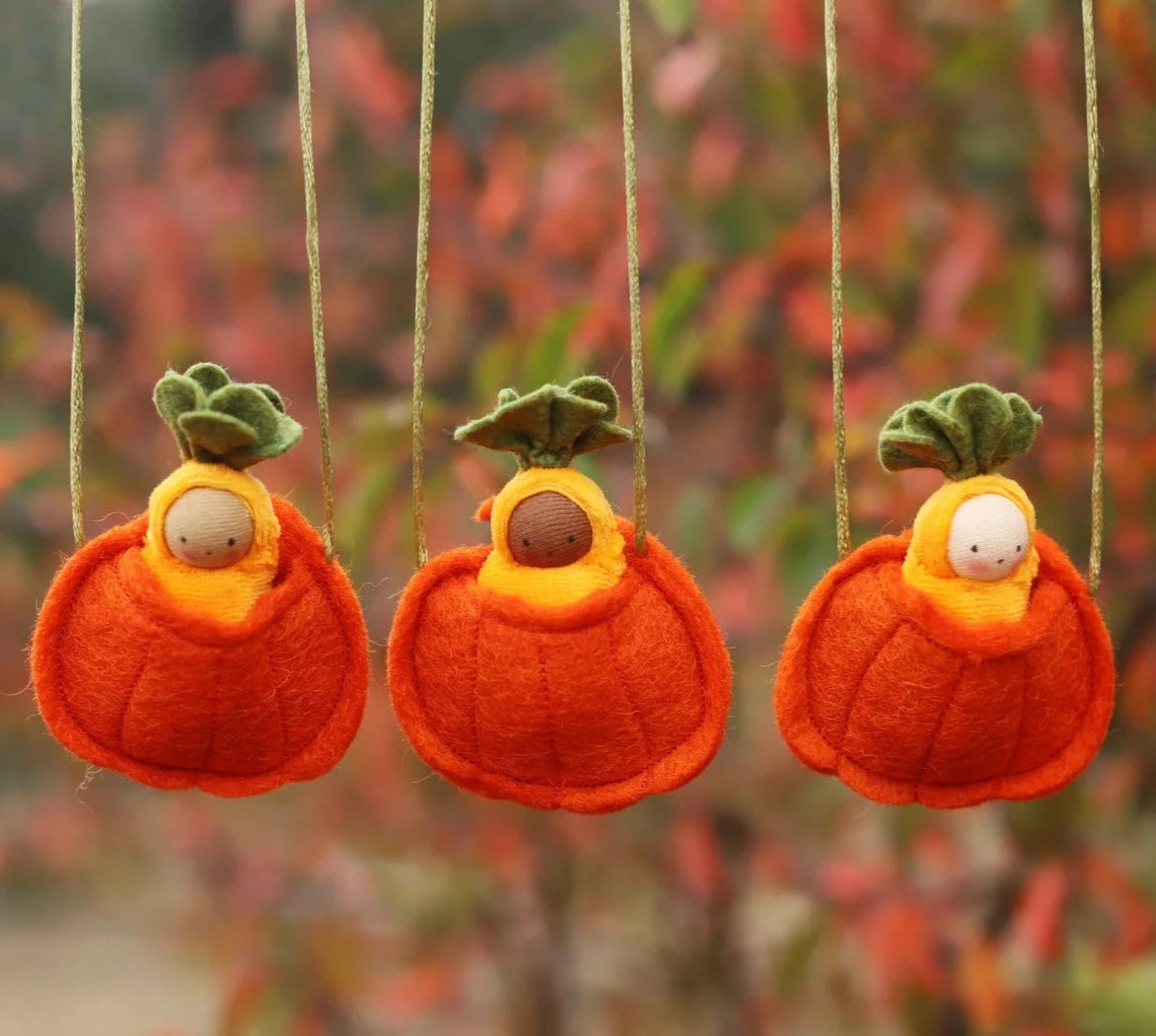 Fairy Shadow Pumpkin Seedling Necklaces, all three skin tone variants hanging side by side outdoors against an autumn foliage background, showing light, dark, and white-skin knitted seedling dolls in yellow hoods nestled in felted orange wool pumpkin pouches with green felt leaf tops on natural cord straps
