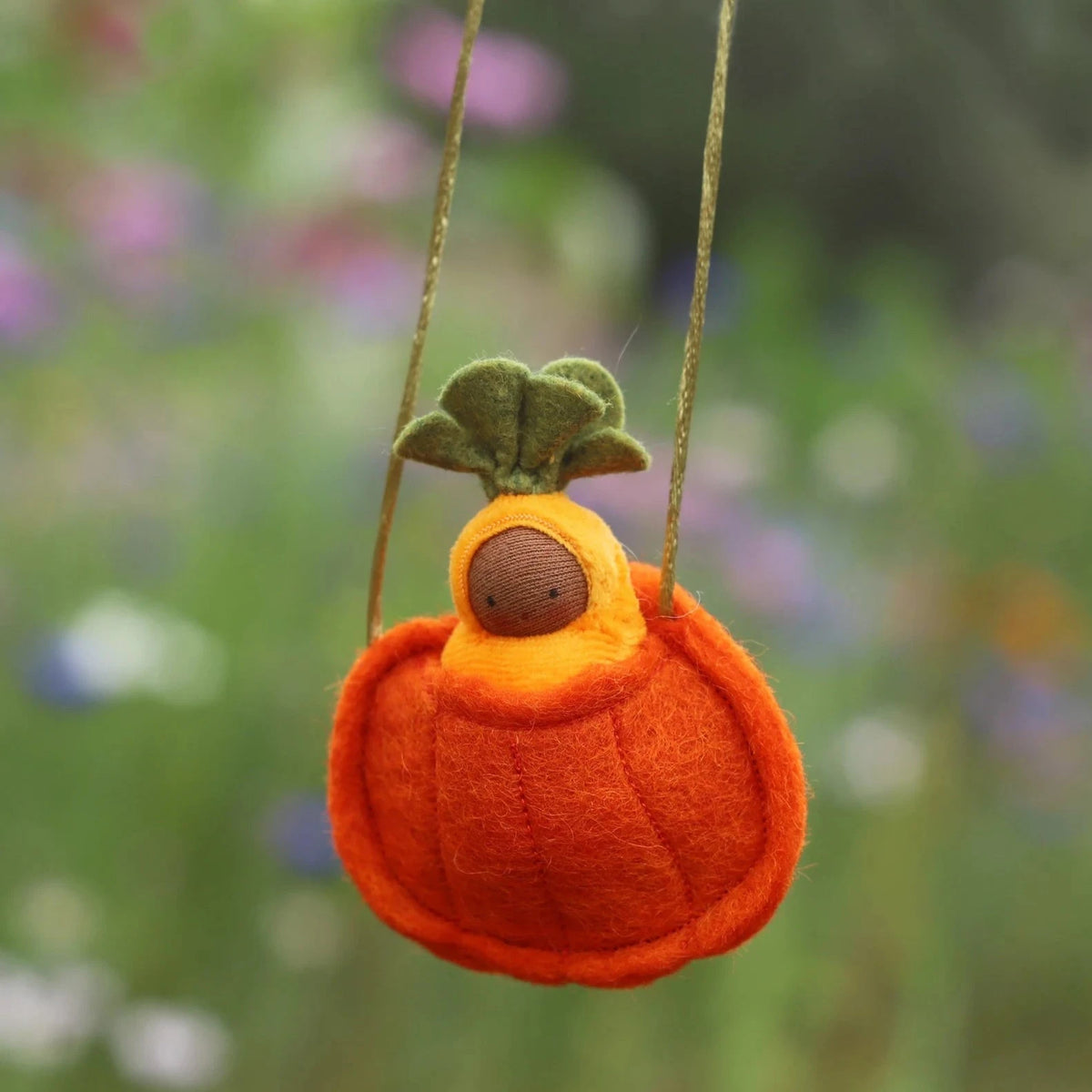 Fairy Shadow Pumpkin Seedling Necklace in dark skin tone, close-up hanging against a soft floral bokeh background, showing a knitted dark-skin seedling doll in a yellow hood nestled in a felted orange ribbed pumpkin pouch with green felt leaf top on a gold metallic cord

