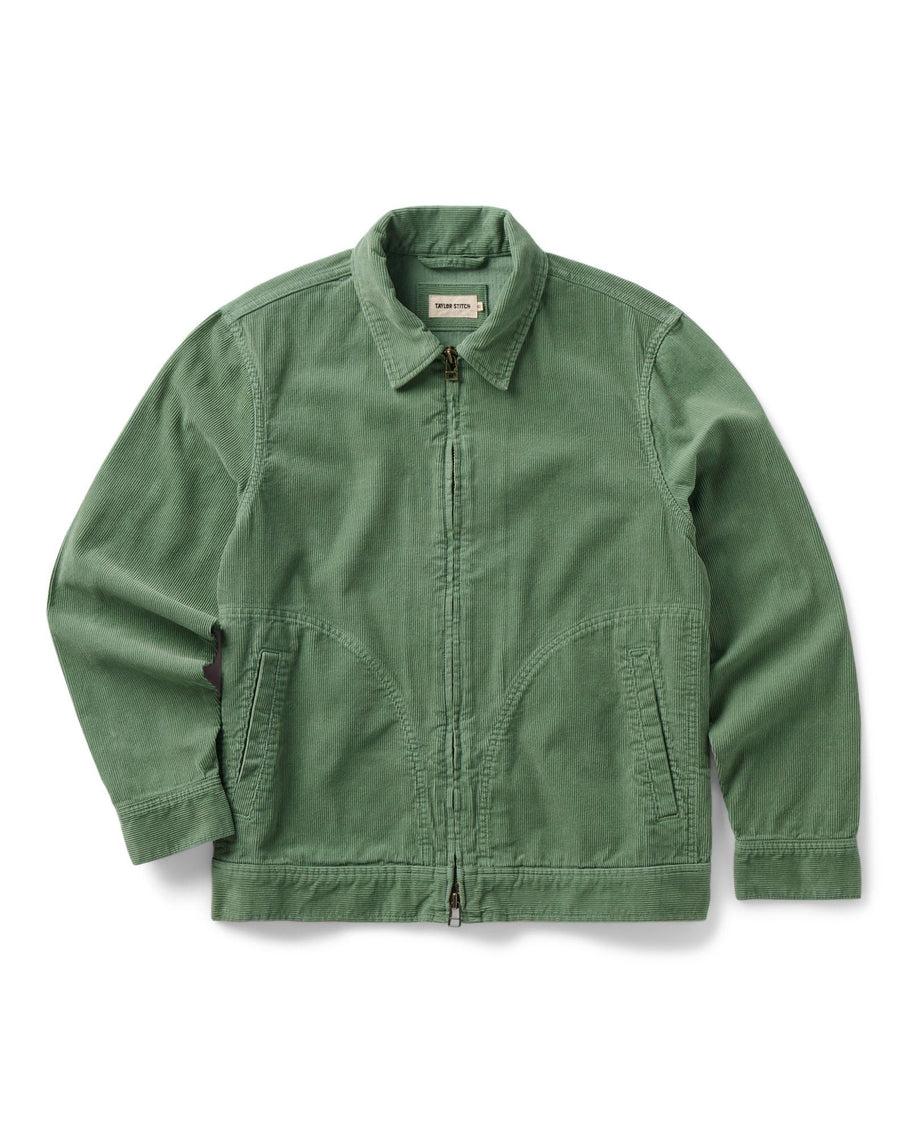 Flat lay of the Taylor Stitch Piston Jacket in Ivy Corduroy on a white background, showing the full jacket from the front. The jacket is a muted sage green in wide-wale organic cotton corduroy with a point collar, a YKK two-way separating zipper down the center front, two curved lower hand warmer pockets, long sleeves with single-button cuffs, and a ribbed hem. A small woven Taylor Stitch label is visible at the collar.
