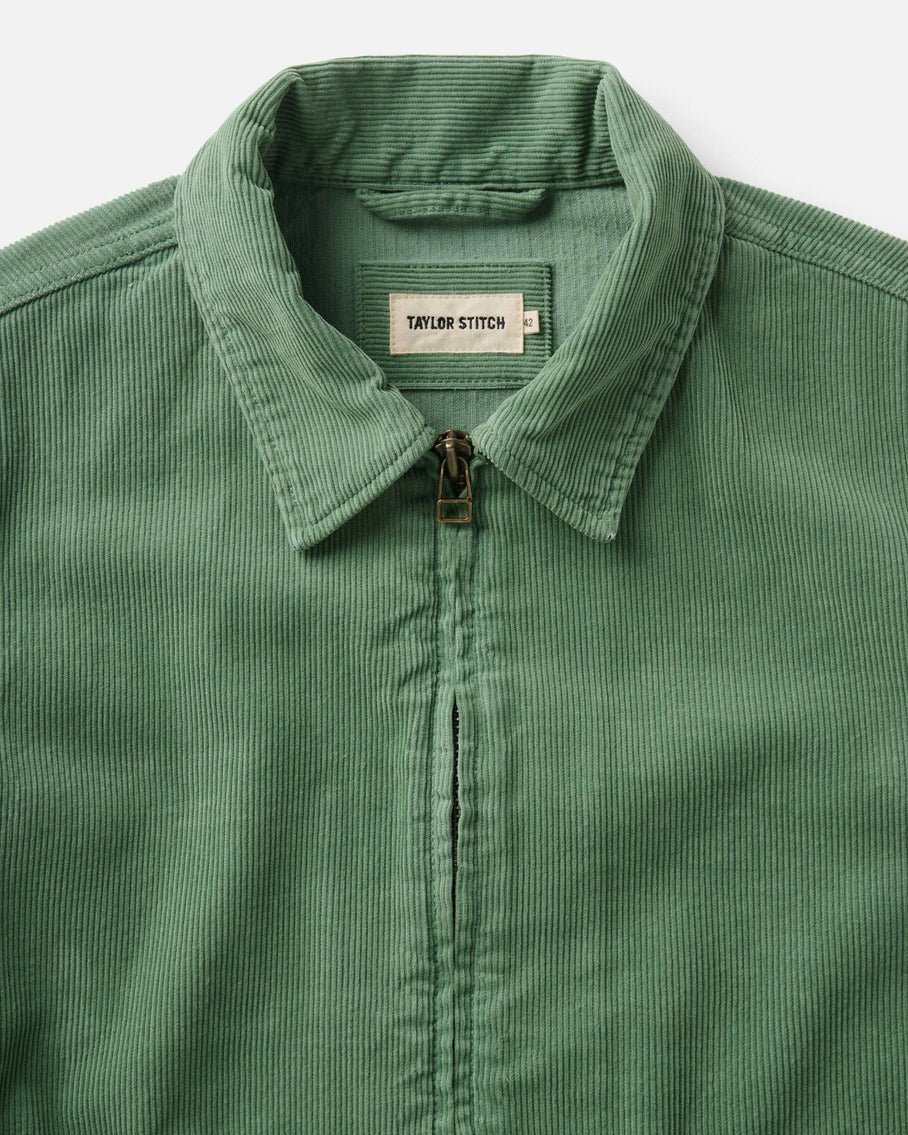 Detail shot of the collar and upper chest of the Taylor Stitch Piston Jacket in Ivy Corduroy, flat lay on a light gray surface. The wide-wale corduroy texture fills the frame in sage green. The woven Taylor Stitch label in cream with black lettering is centered at the back collar. The antique brass YKK zipper pull is visible at center front, with the zipper teeth running down from the collar.
