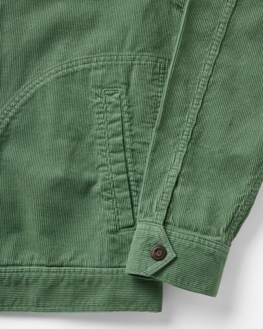 Detail shot of the lower left body and cuff of the Taylor Stitch Piston Jacket in Ivy Corduroy on a light gray surface, showing the wide-wale organic cotton corduroy texture up close. The curved hand warmer pocket opening and zip placket panel are visible at left, with double-needle felled seam construction throughout. The single-button cuff tab at the sleeve hem features a dark gunmetal snap button, and the ribbed hem band runs along the bottom edge.
